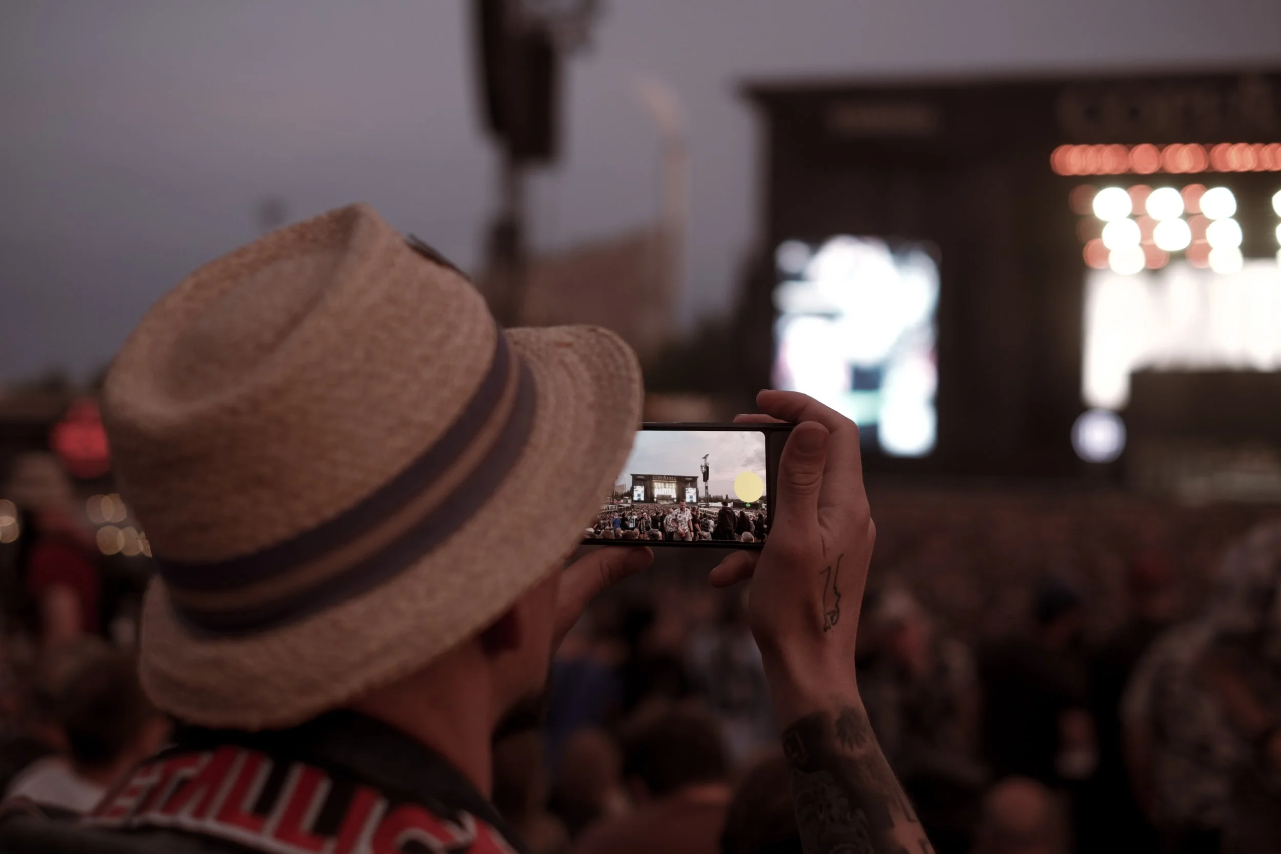 Person med stråhat, der tager et billede med en smartphone af en scene på en koncert eller festival, med mange folk i publikum.