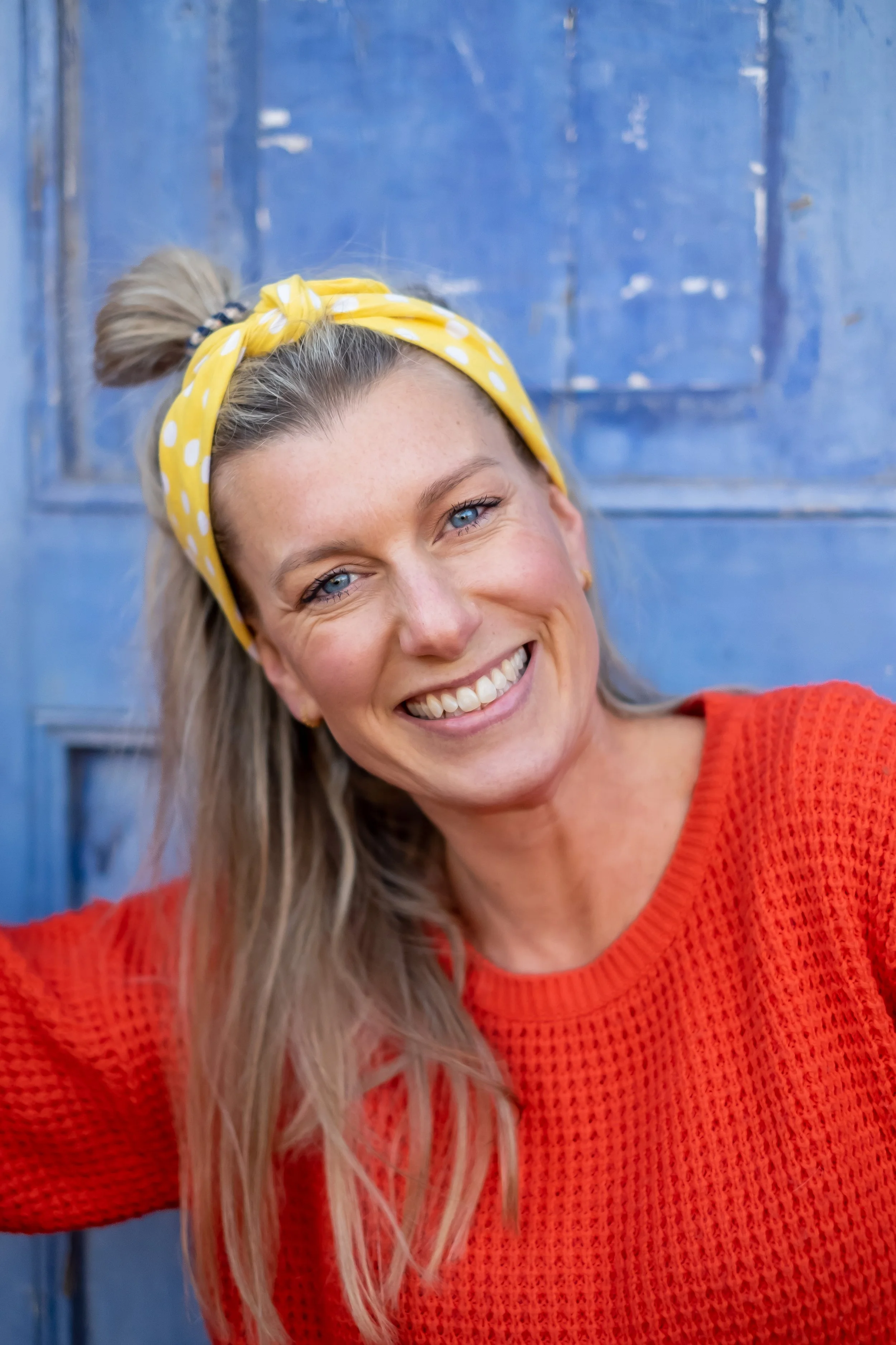 En smilende kvinde med blondt hår, iført en rød sweater og en gul, polka-dotted hårbånd, foran en blå dør.