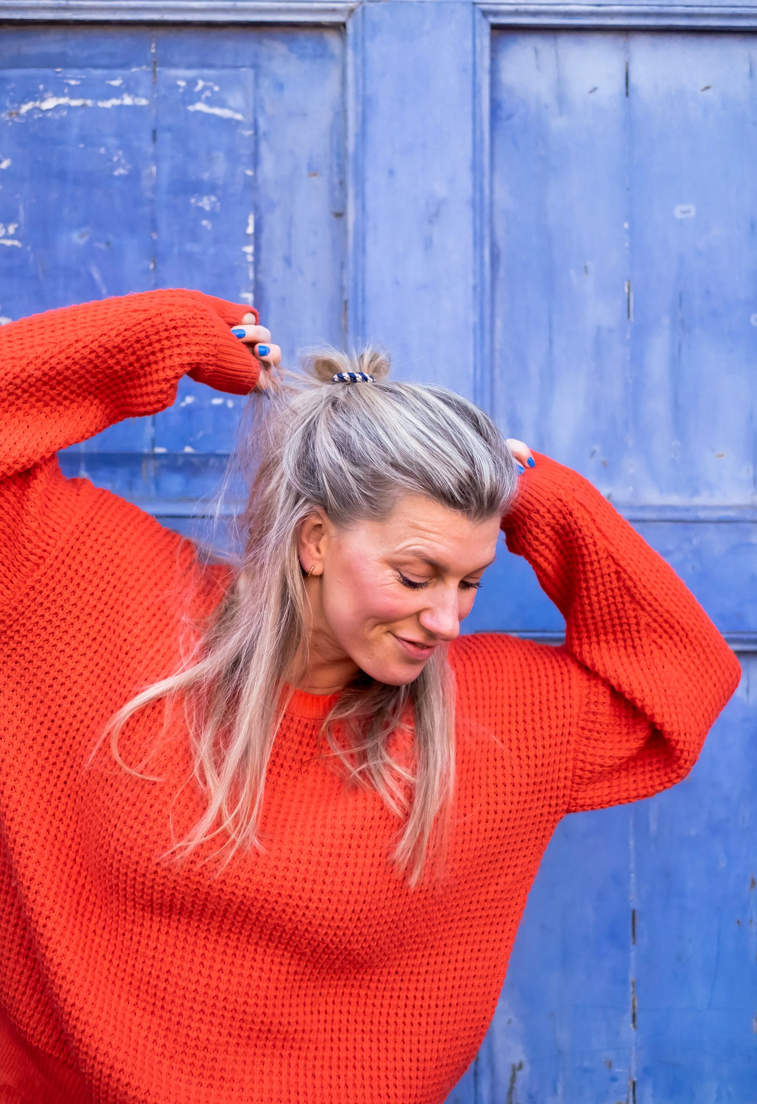 En kvinde med gråt hår i en rød, strikket sweater, løfter sit hår op med hænderne foran en blå dør.