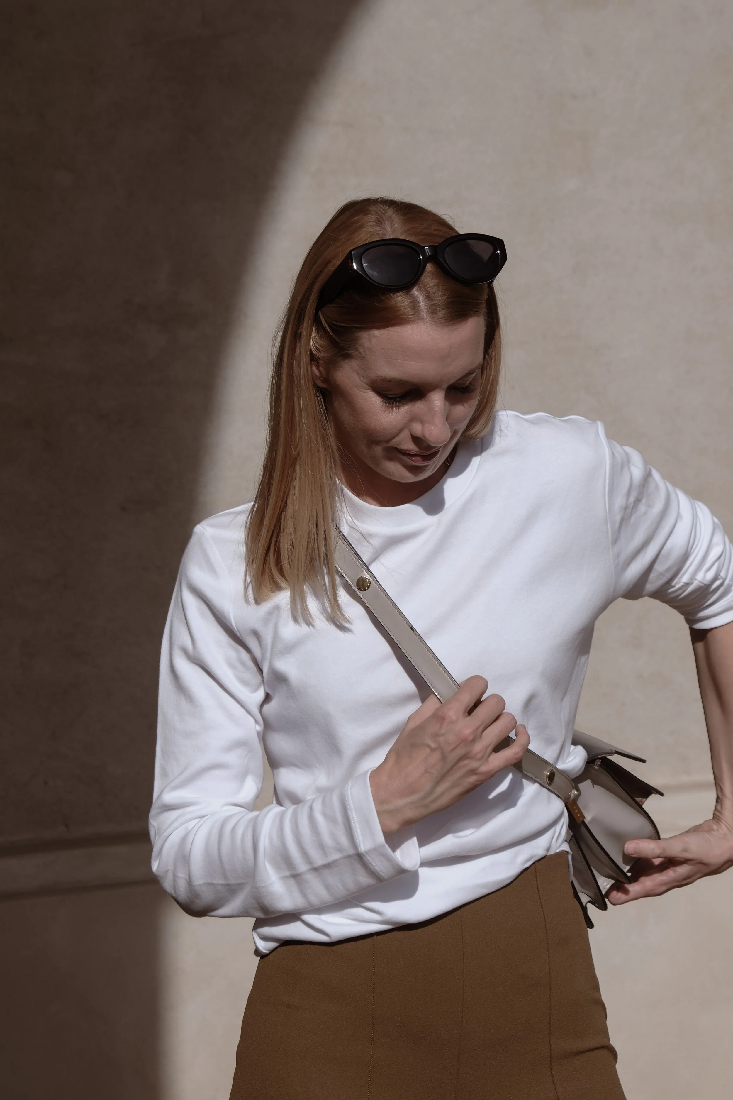 Kvindelig person iført hvid langærmet trøje, brunt skjørt, med solbriller på hovedet, står mod venstre og ser ned. Bærer en beige taske over skulderen, er ved at tage den af, med en neutral beige baggrund.