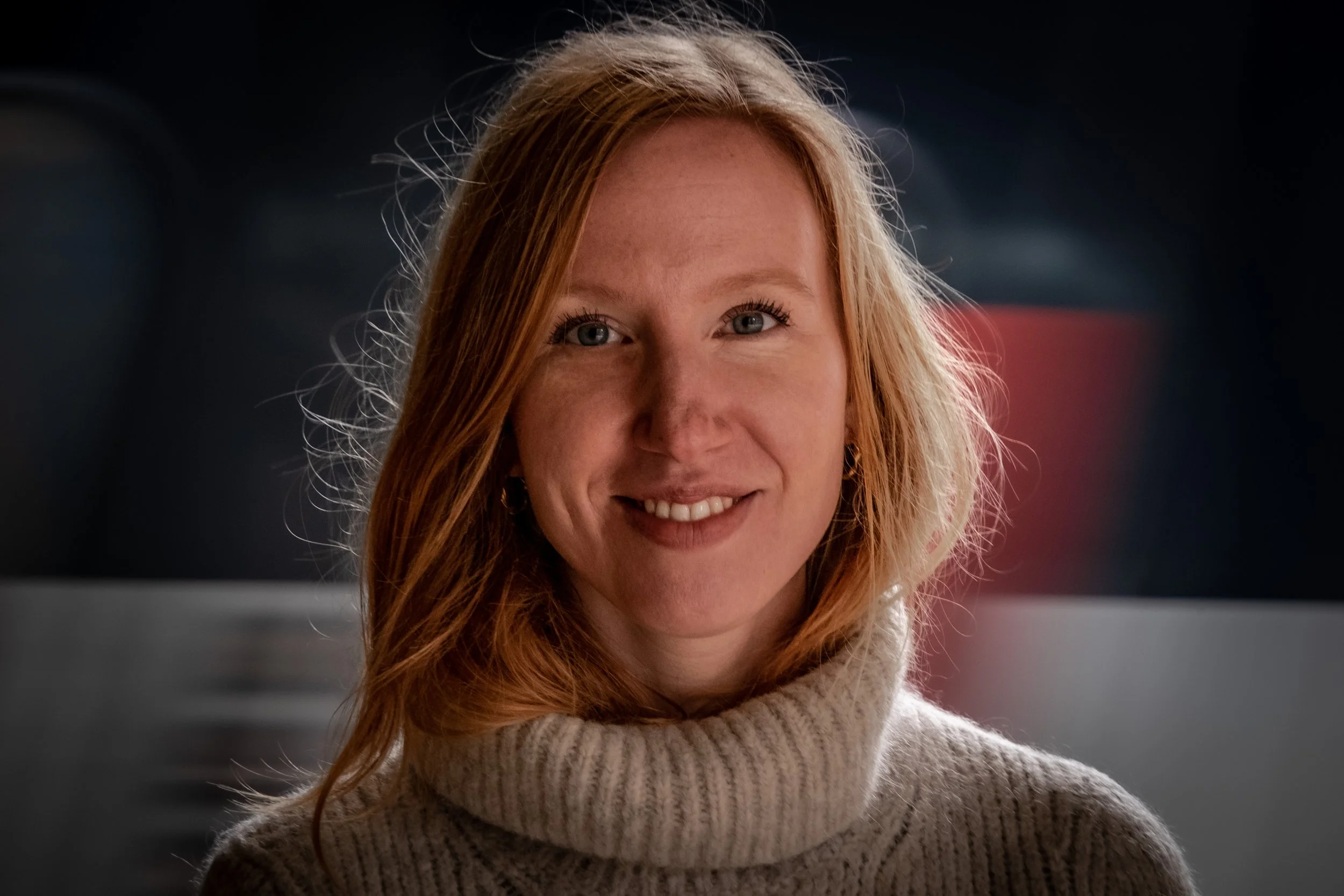 Et portrait af en ung kvinde med rødt hår og en beige striktrøje, der smiler, foran en mørk baggrund.