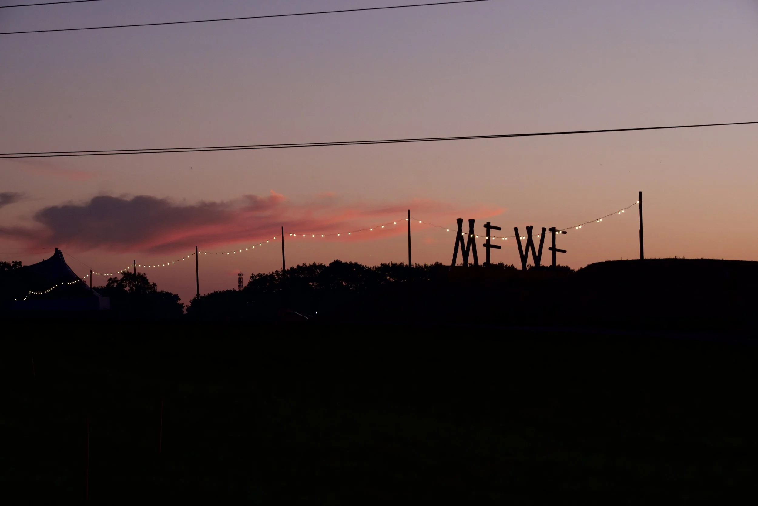 A sunset sky with pink and purple clouds, string lights, and a sign spelling "ME WE" in the distance under a darkening horizon.