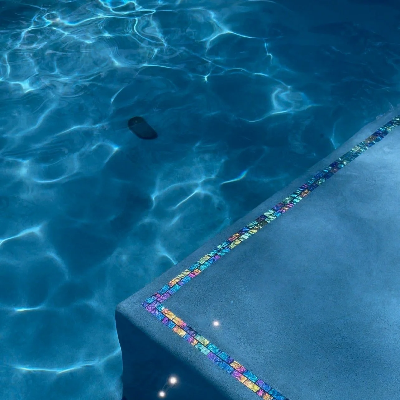 Swimming pool with colorful mosaic tiles on the edge and submerged dark object.