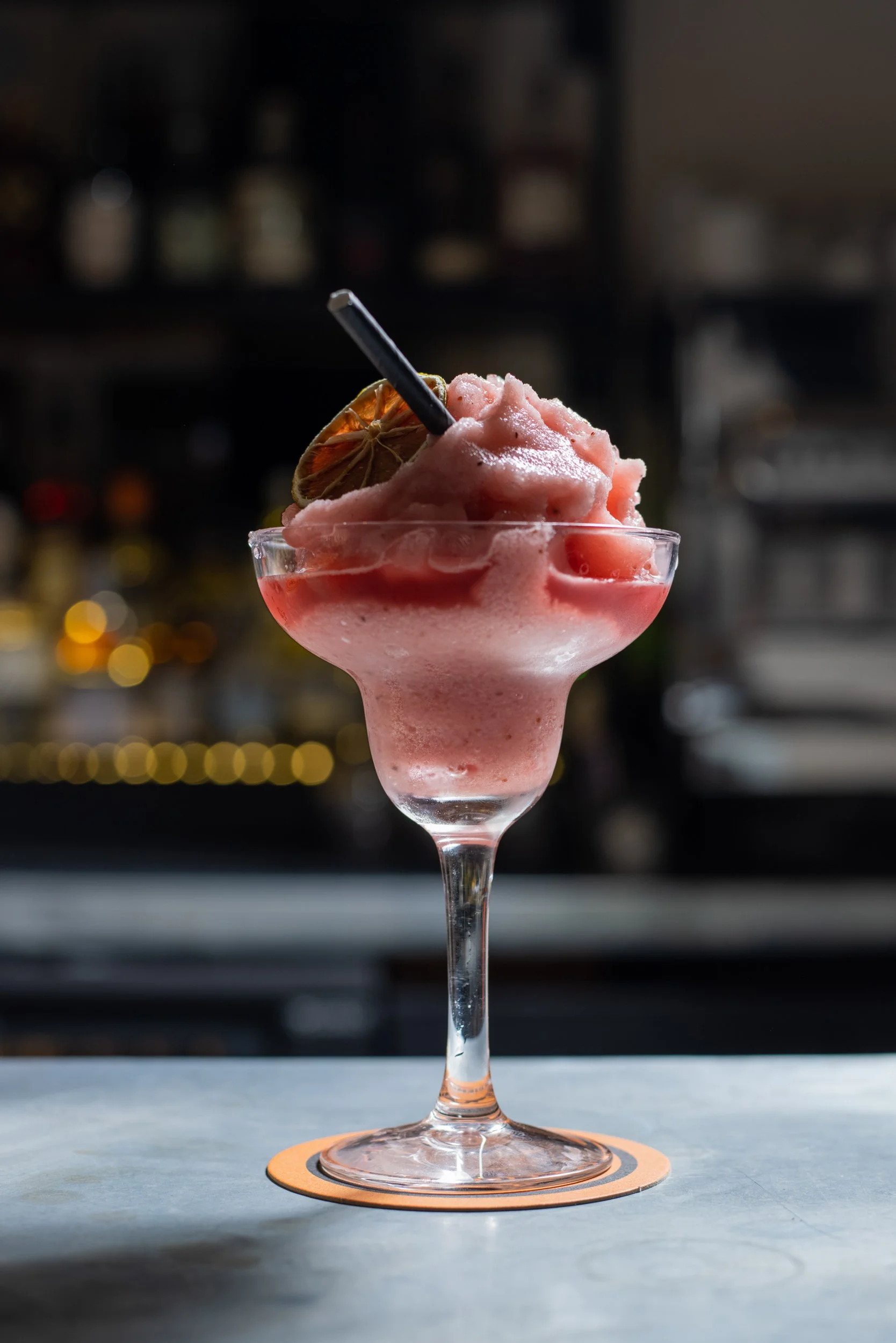 Pink frozen cocktail in a glass with a straw and a dried lime slice on top, placed on a coaster on a countertop with blurred background.