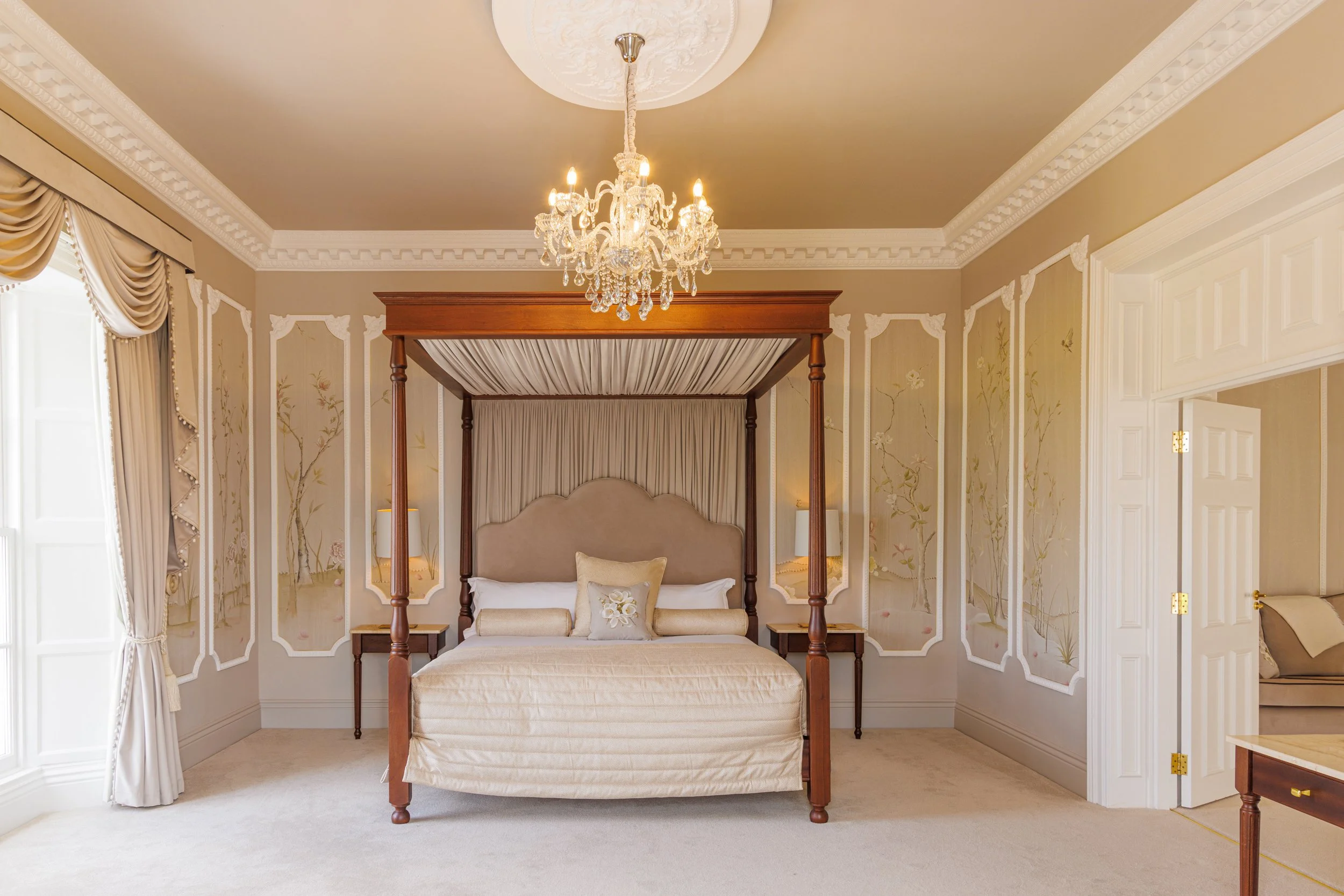 Luxurious bedroom with a chandelier, four-poster bed with beige bedding, and elegant draped curtains.