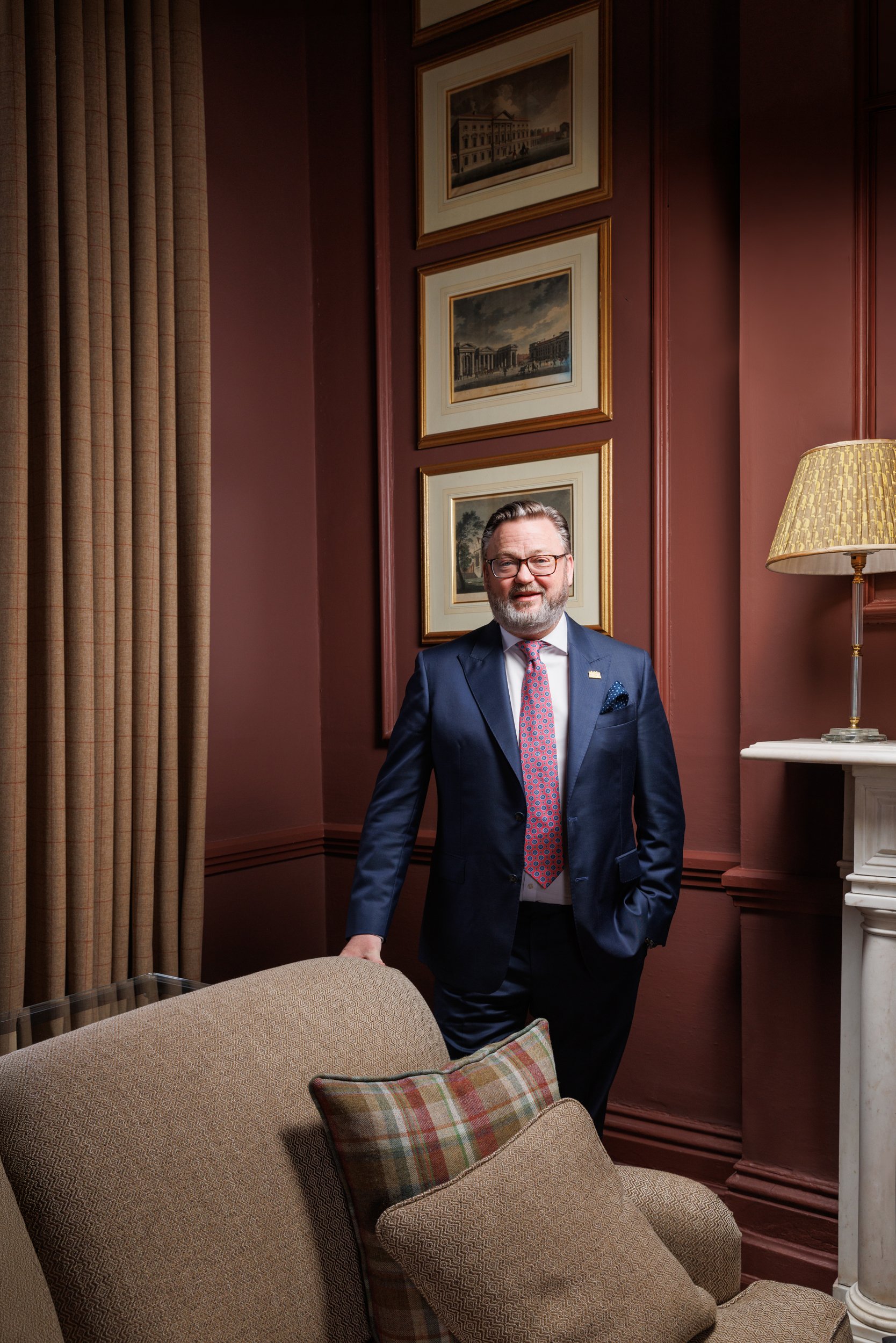 A man in a blue suit, glasses, and a pink patterned tie standing in a room with burgundy walls, framed artwork, a beige sofa with plaid pillows, a lamp, and thick curtains.