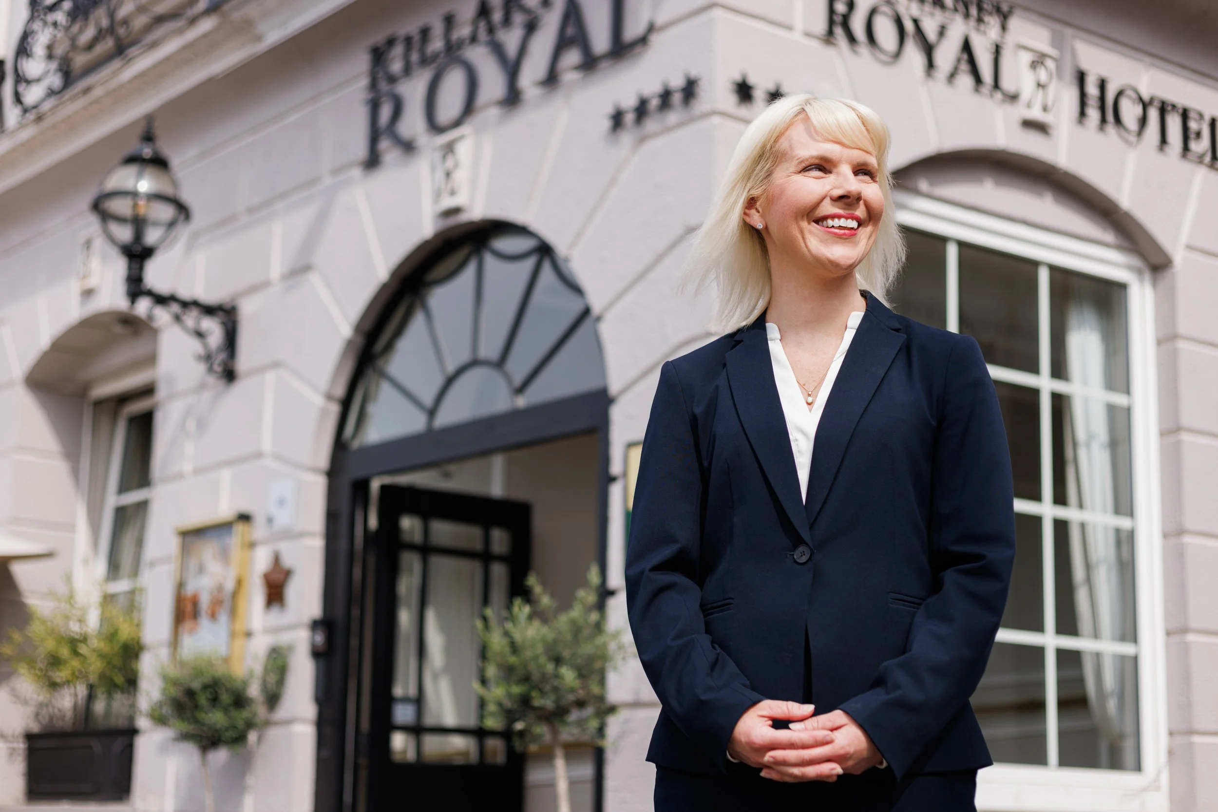 A woman in a navy blazer and white blouse smiling outside a building with a sign that reads "Royal Hotel".