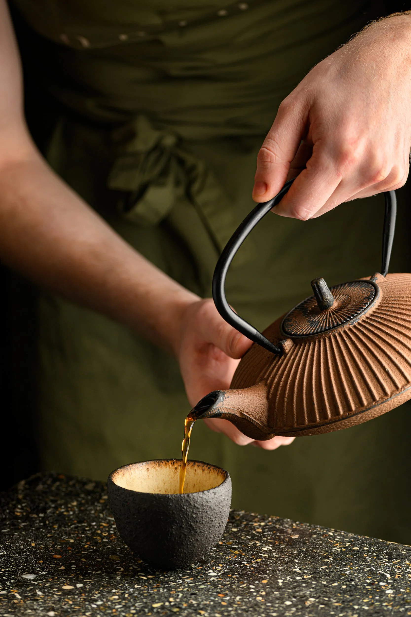 A person pouring tea from a brown teapot into a black cup on a speckled countertop.