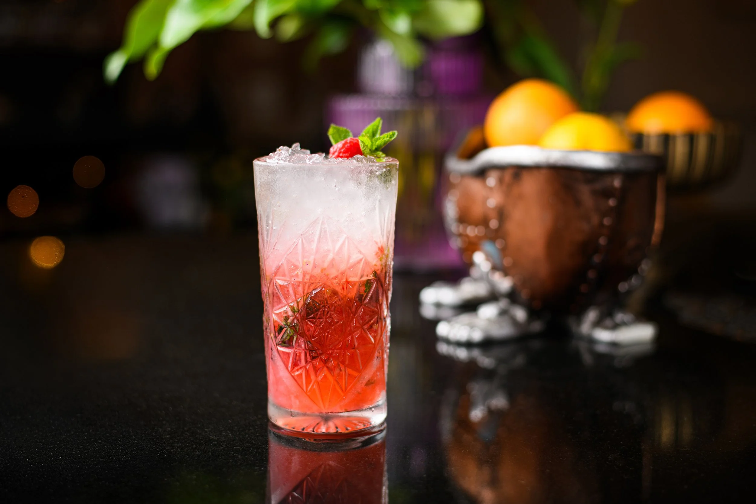 A pink cocktail in a tall, textured glass garnished with a raspberry and a mint sprig, placed on a dark reflective surface with a blurred background featuring oranges and a copper container.