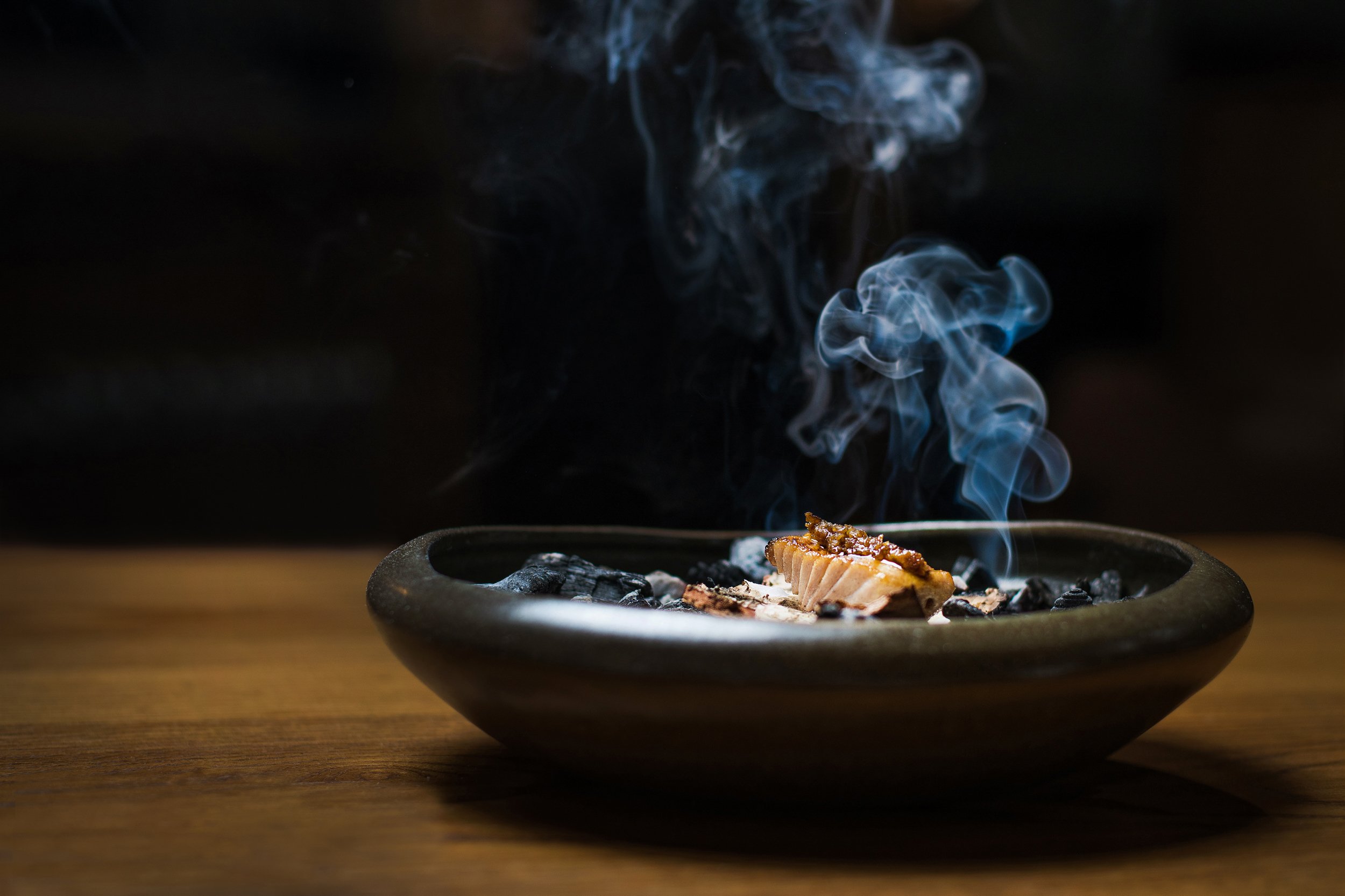 A small smoking fire pit on a wooden surface with a piece of meat or fish on top, emitting blue smoke against a dark background.