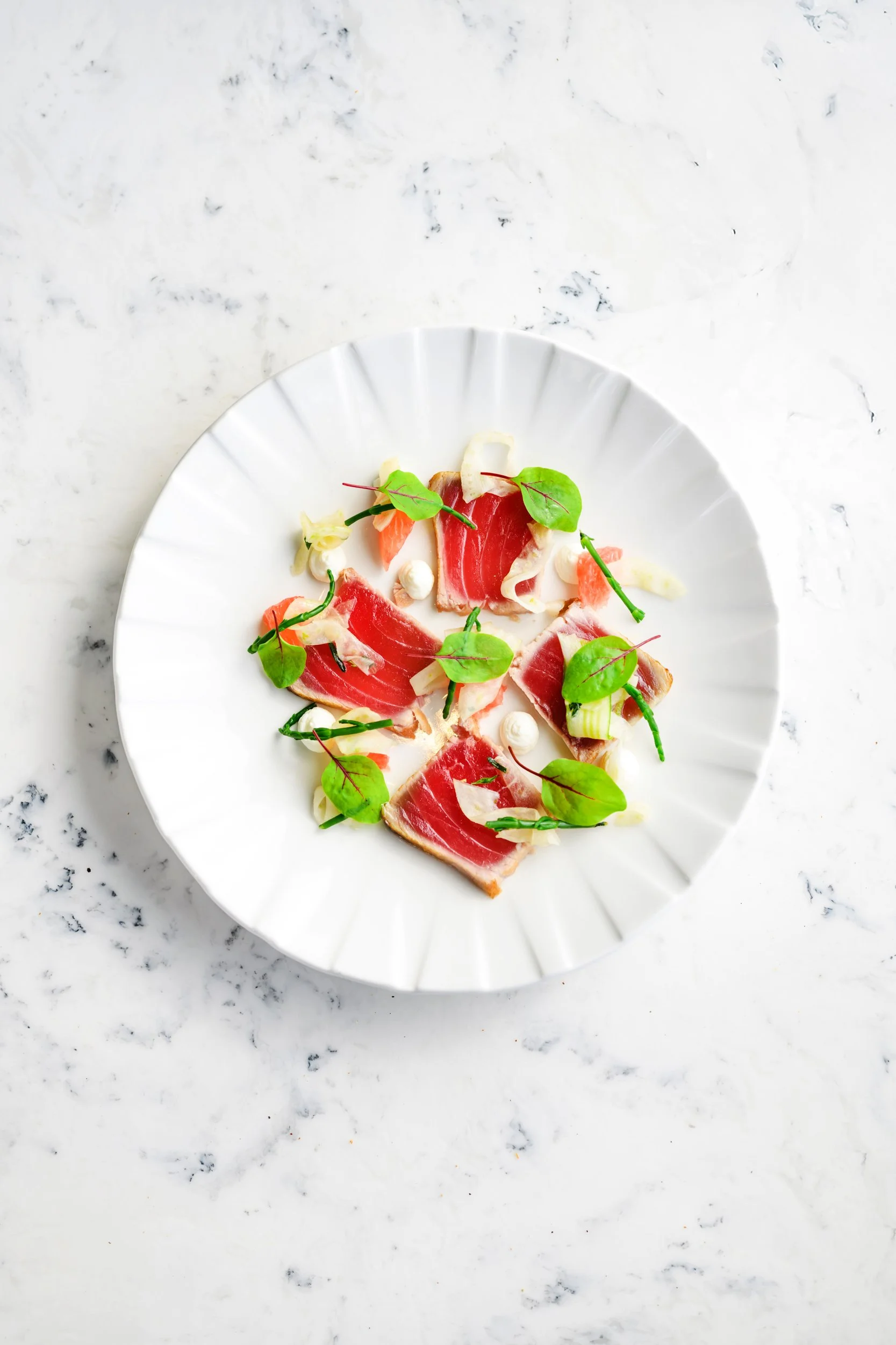 Sliced prosciutto on a white plate with green microgreens and small dollops of white creamy sauce, on a white marble surface.