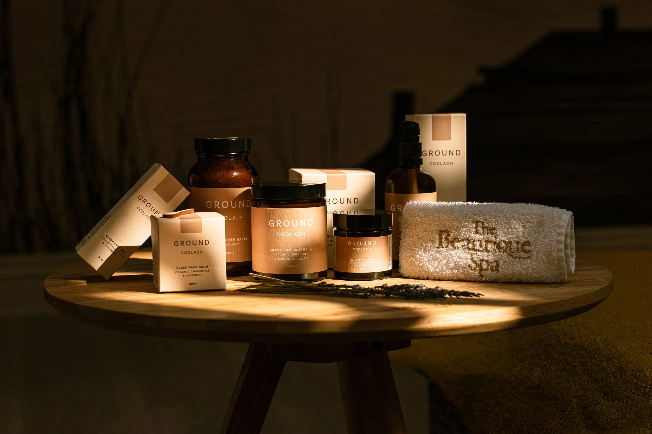 Spa products, including creams, balms, and oils, are displayed on a wooden table with a rolled towel embroidered with "The Beauteque Spa" in a dimly lit setting.