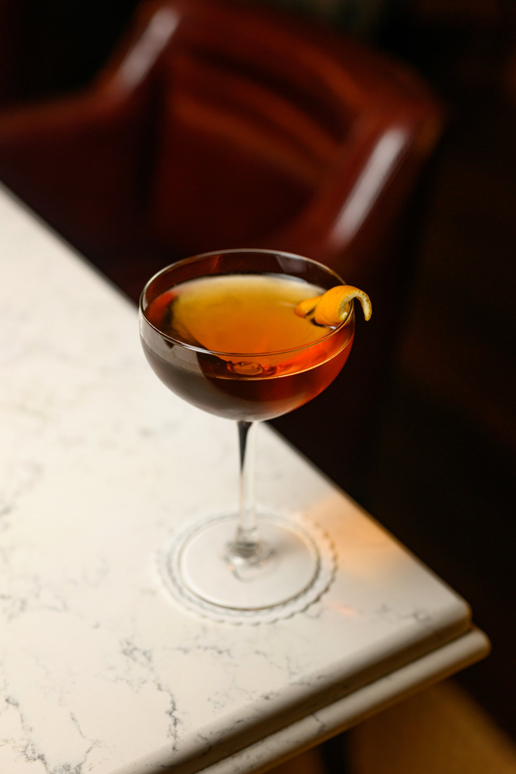 A cocktail glass filled with a dark red drink, garnished with a lemon twist, placed on a white marble surface with black veining.