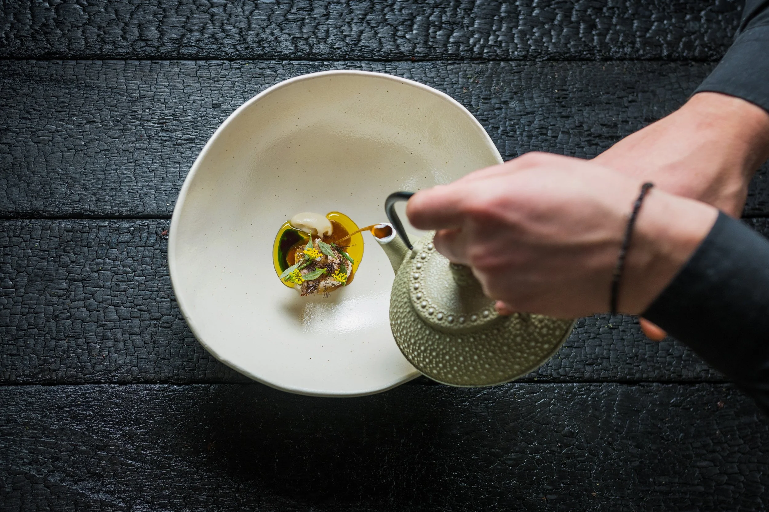 A person holding a teapot pouring a drink into a small bowl on a white plate, on a black textured wooden surface.