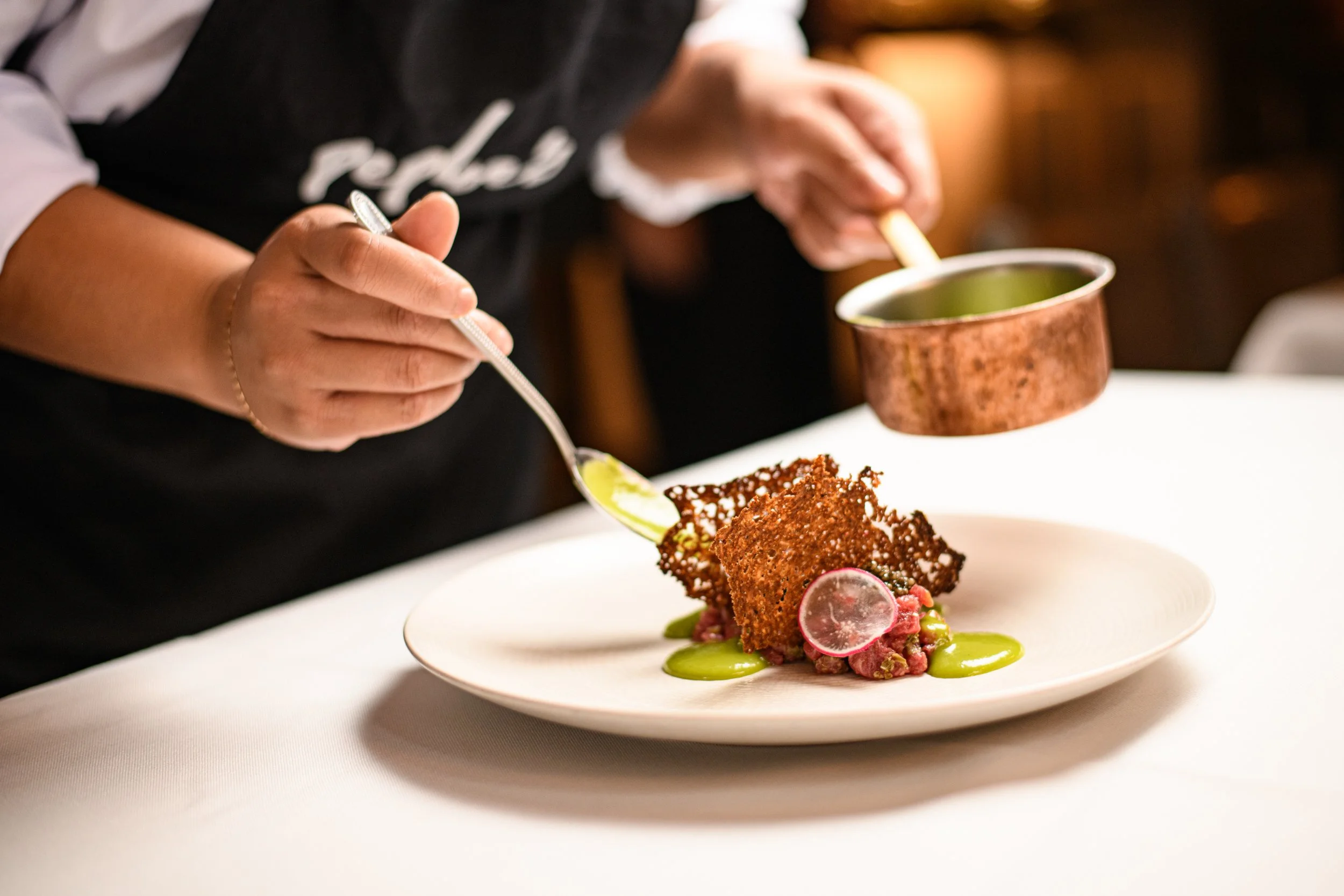 A person in a black apron is plating a dessert with a spoon; the dessert includes a crispy, caramelized sugar piece, pink radish slices, and green sauces on a white plate.