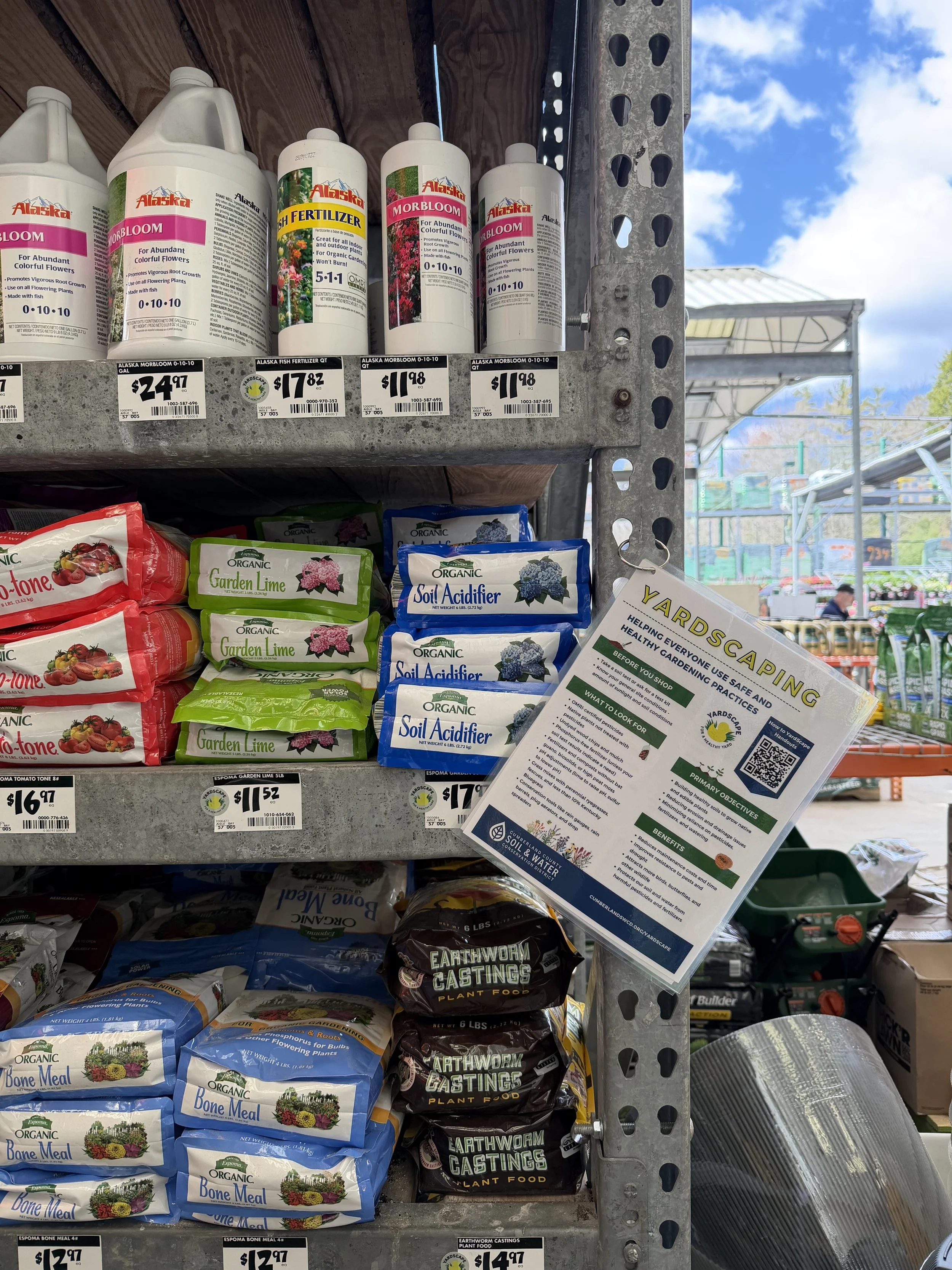 Shelves stocked with bags of organic soil amendment products, labeled 'Bone Meal,' 'Soil Acidifier,' 'Garden Lime,' and 'Earthworm Castings.' A sign with gardening information is visible in front of the shelves, and the sky can be seen through the open side of the store.
