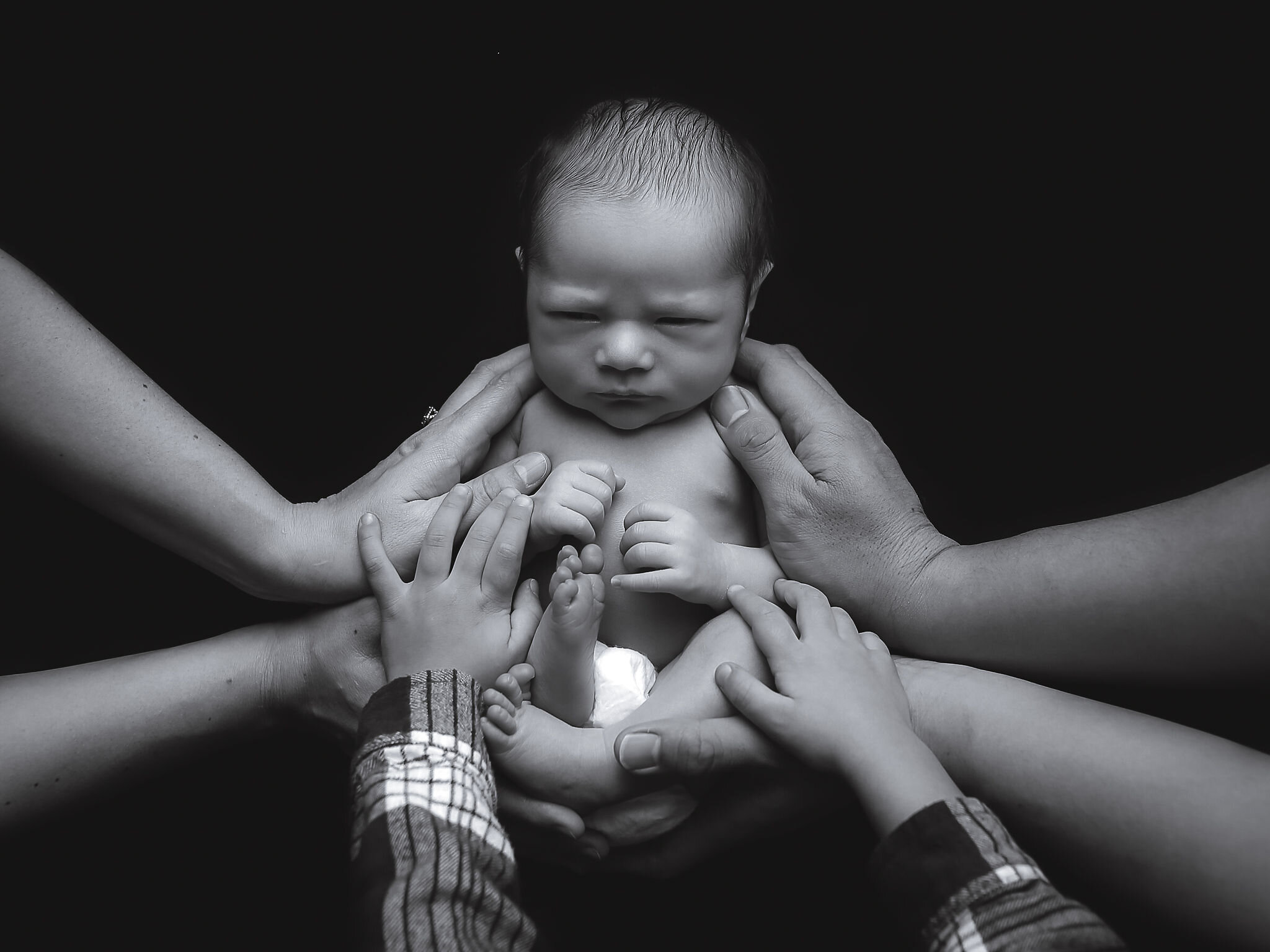 black and white image of a newborn baby being held by 6 family hands