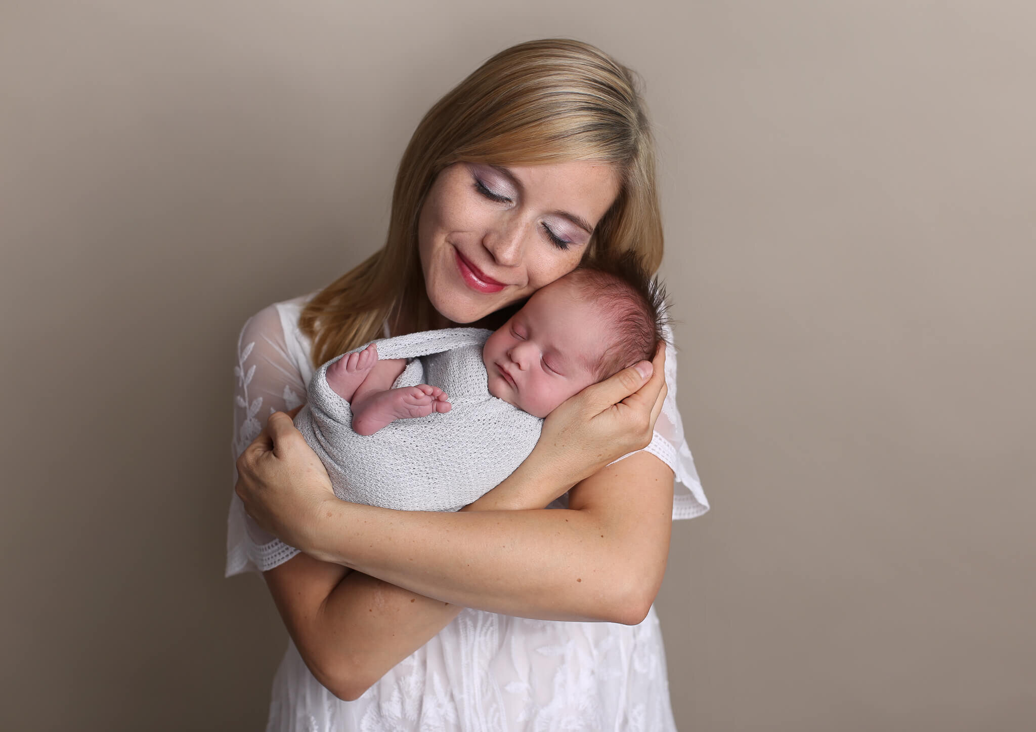  A picture of a new mother cradling her newborn baby in her arms, leaning her cheek softly on his head as he lies wrapped up, sleeping with his tiny feet showing by Photography by L Rose - San Diego CA newborn photography 