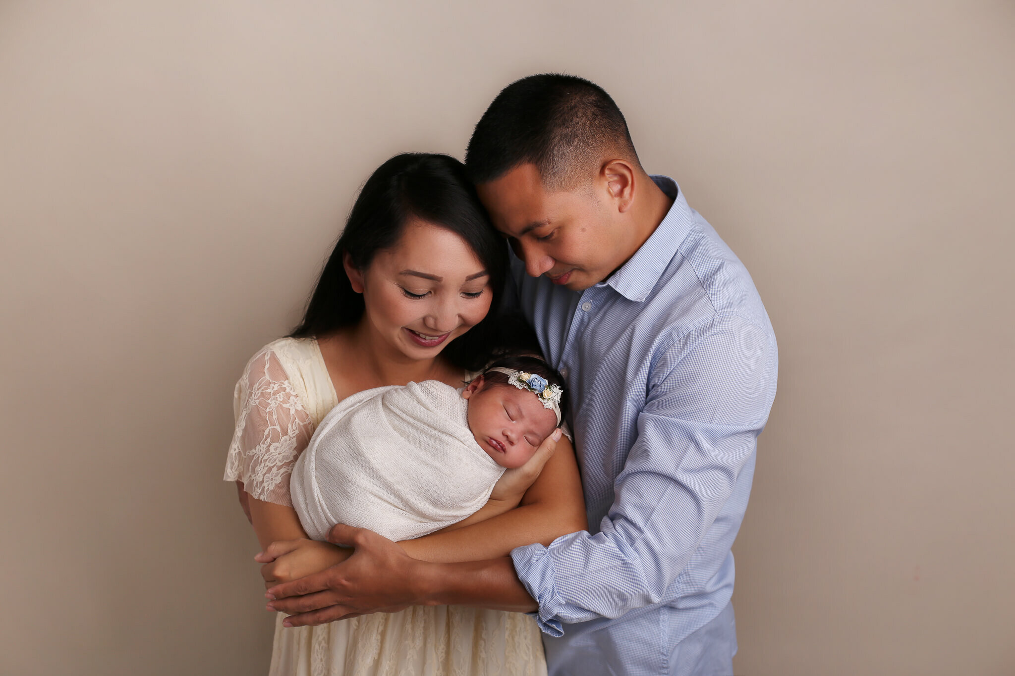  An image of a new family with dad hugging them, touching his head on his mom’s hair, as she holds their newborn baby in her arms from a newborn photo session 