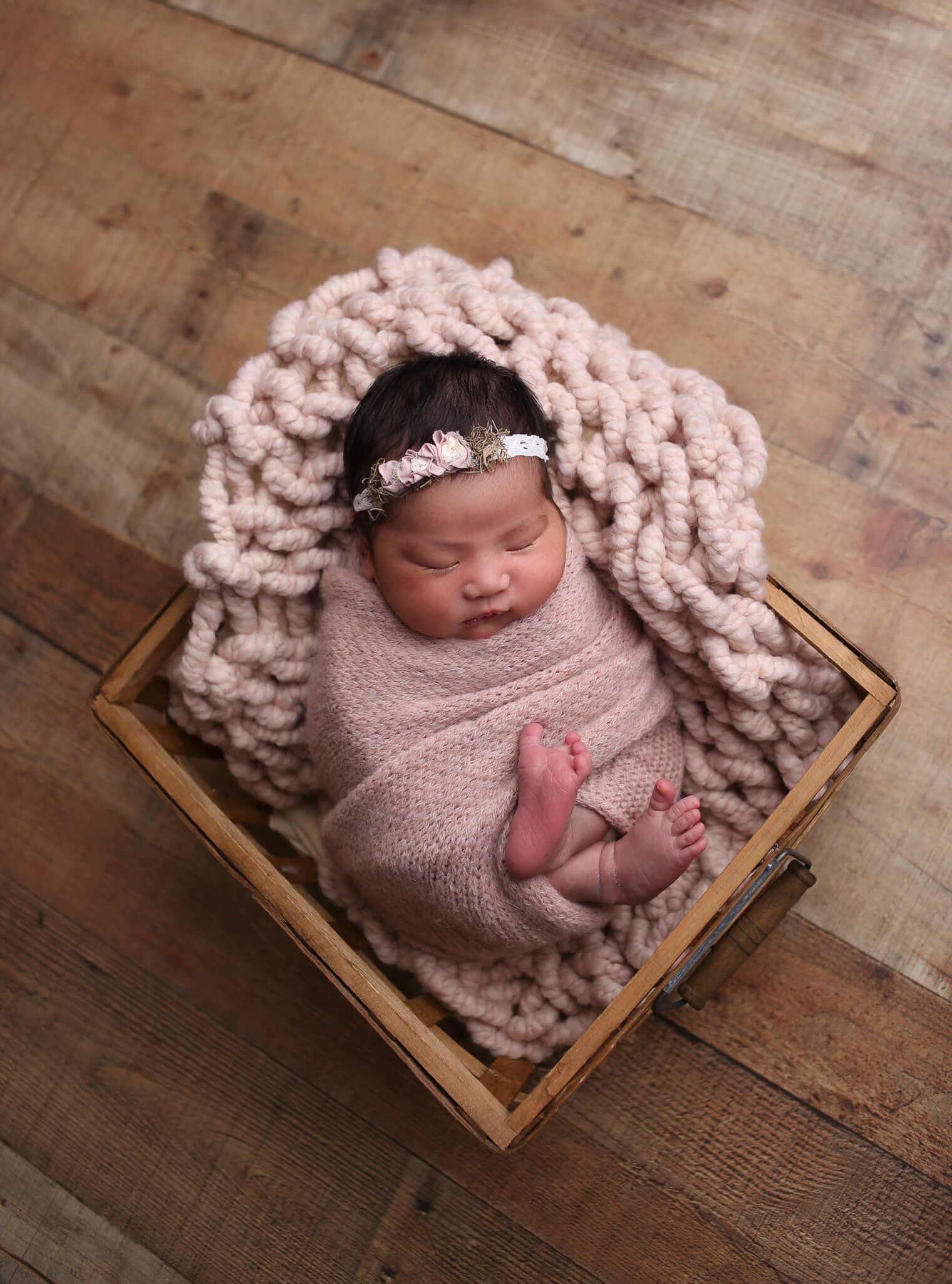  A photograph of a newborn girl in her headband, wrapped in a pink blanket with her tiny feet showing, sleeping in a wooden basket from a newborn picture session 