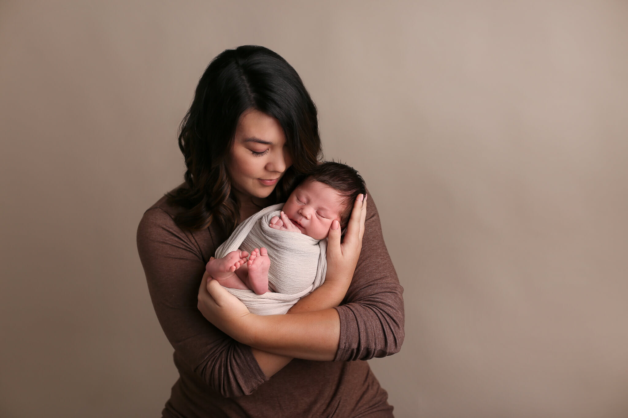  A picture of a new mother gazing down at her newborn child wrapped in gauze, cradled in her arms as his little hands and feet peek out from a baby photo session 