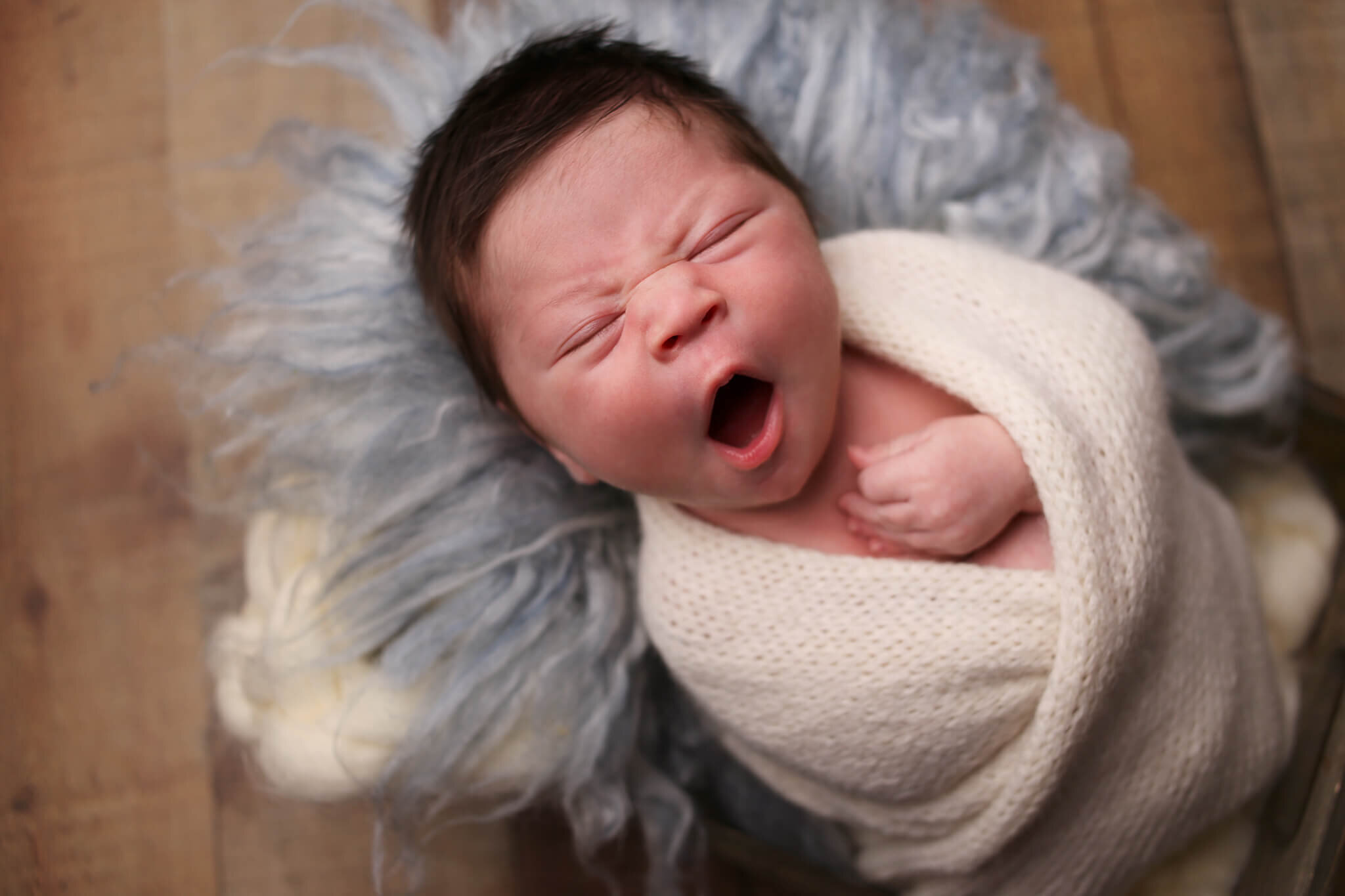  A photograph of a newborn baby with a big yawn, wrapped up in a blanket with its head resting on a soft bed of yarn by Photography by L Rose - San Diego California newborn photographer 