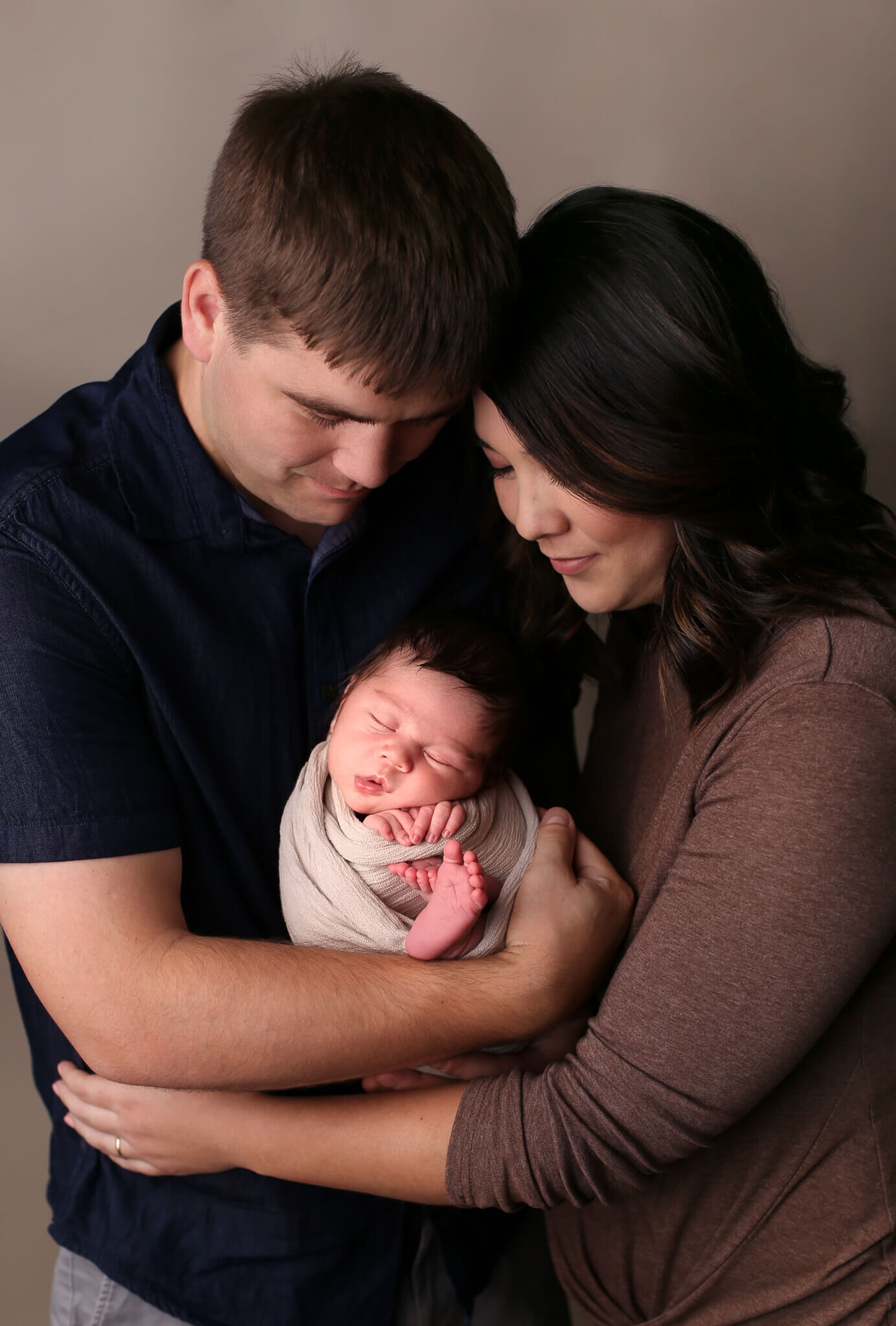  An image of a father and mother standing close, gazing down as dad holds the sleeping newborn baby with its tiny hands and feet showing through the blanket by Photography by L Rose - San Diego newborn photography 