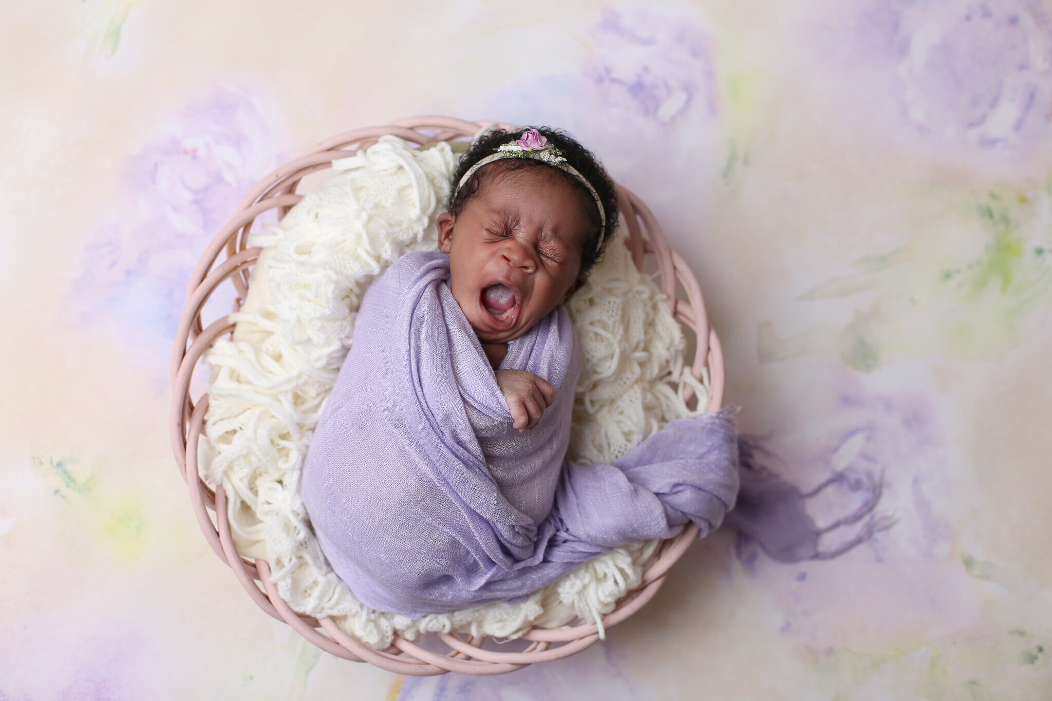  An image of a newborn baby in her flowered headband, having a big yawn as she lies tightly wrapped in gauze on top of a soft cushion of yarn in a basket from a newborn photo session 