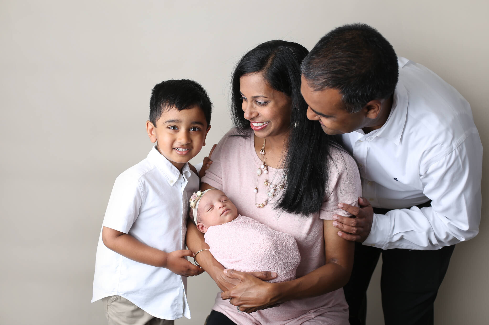  An image of a family grouped close together-with dad leaning over mom’s shoulder, watching the tiny newborn in her arms as big brother stands close by Photography by L Rose - San Diego newborn photography 