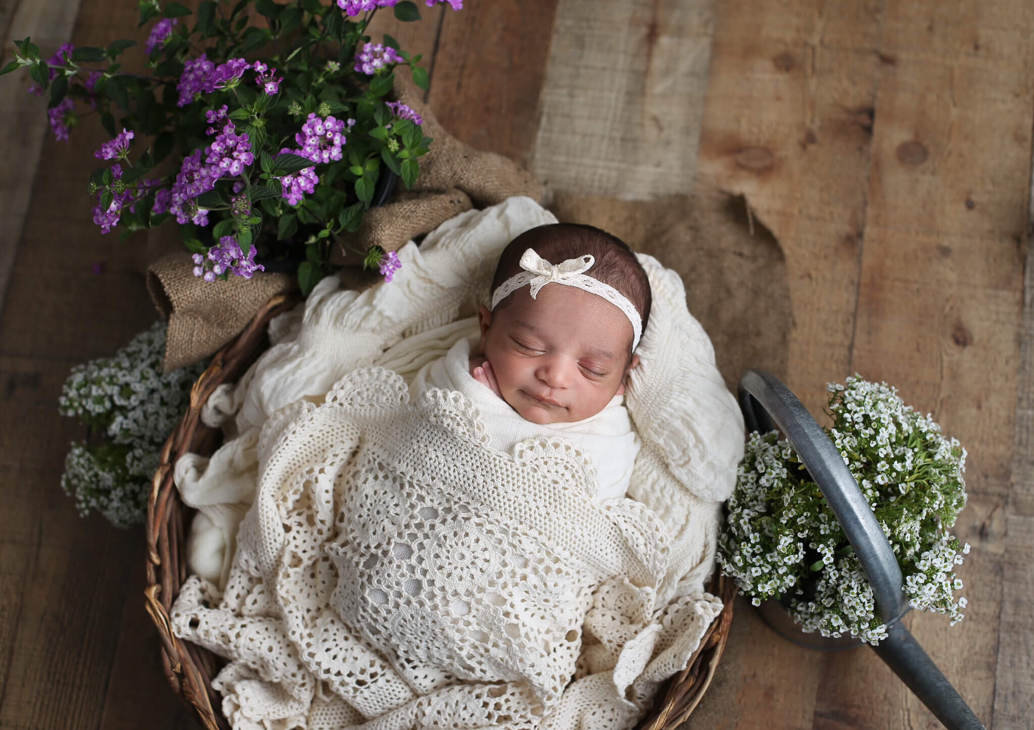  An image of a tiny newborn girl in her hair ribbon, sleeping in a dainty crocheted blanket, surrounded by beautiful flower arrangements from a newborn photo session 