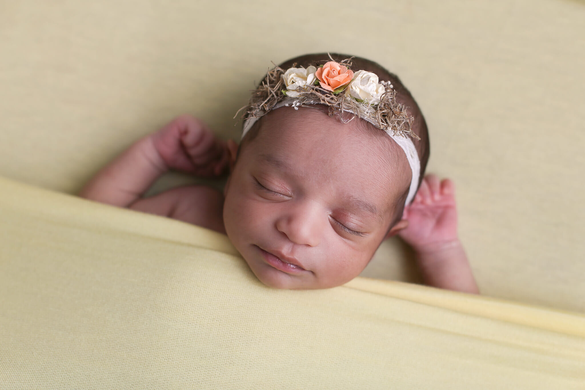  A photograph of a closeup image of a newborn girl wearing a flowered headband as she sleeps soundly in a soft blanket with her tiny hands by her head from a newborn picture session 