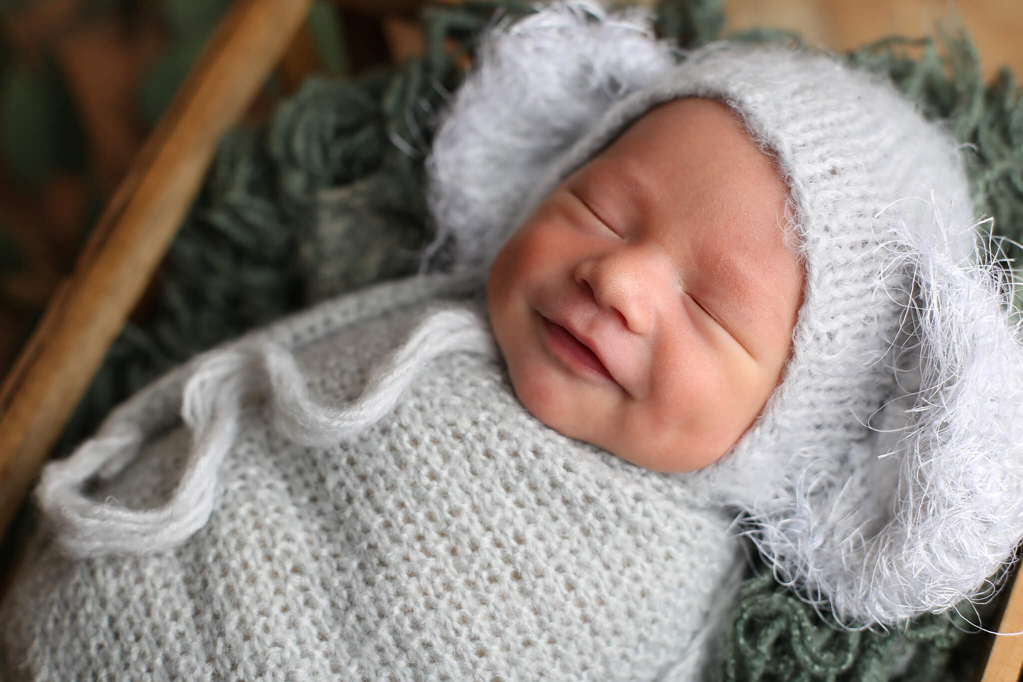  A picture of a new baby asleep in a knitted cap with big fuzzy ears, swaddled in a blanket with a little smile on its face from a baby photo session 