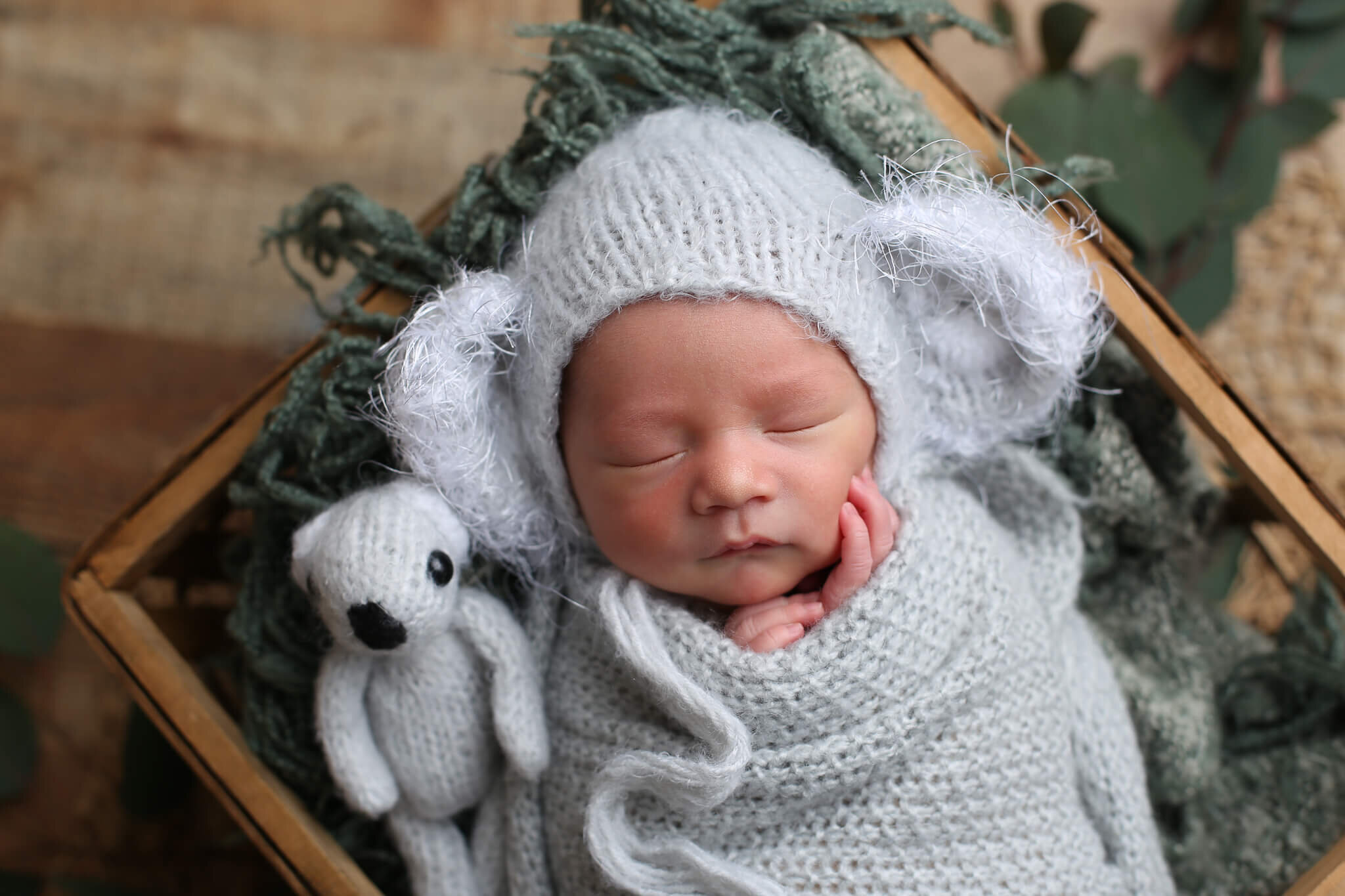  A photograph of a newborn in a knitted cap with big fuzzy ears and wrapped in a blanket with its tiny hands peeking out, sleeping soundly with a stuffed animal, nestled in a wooden basket by Photography by L Rose - San Diego newborn photography 