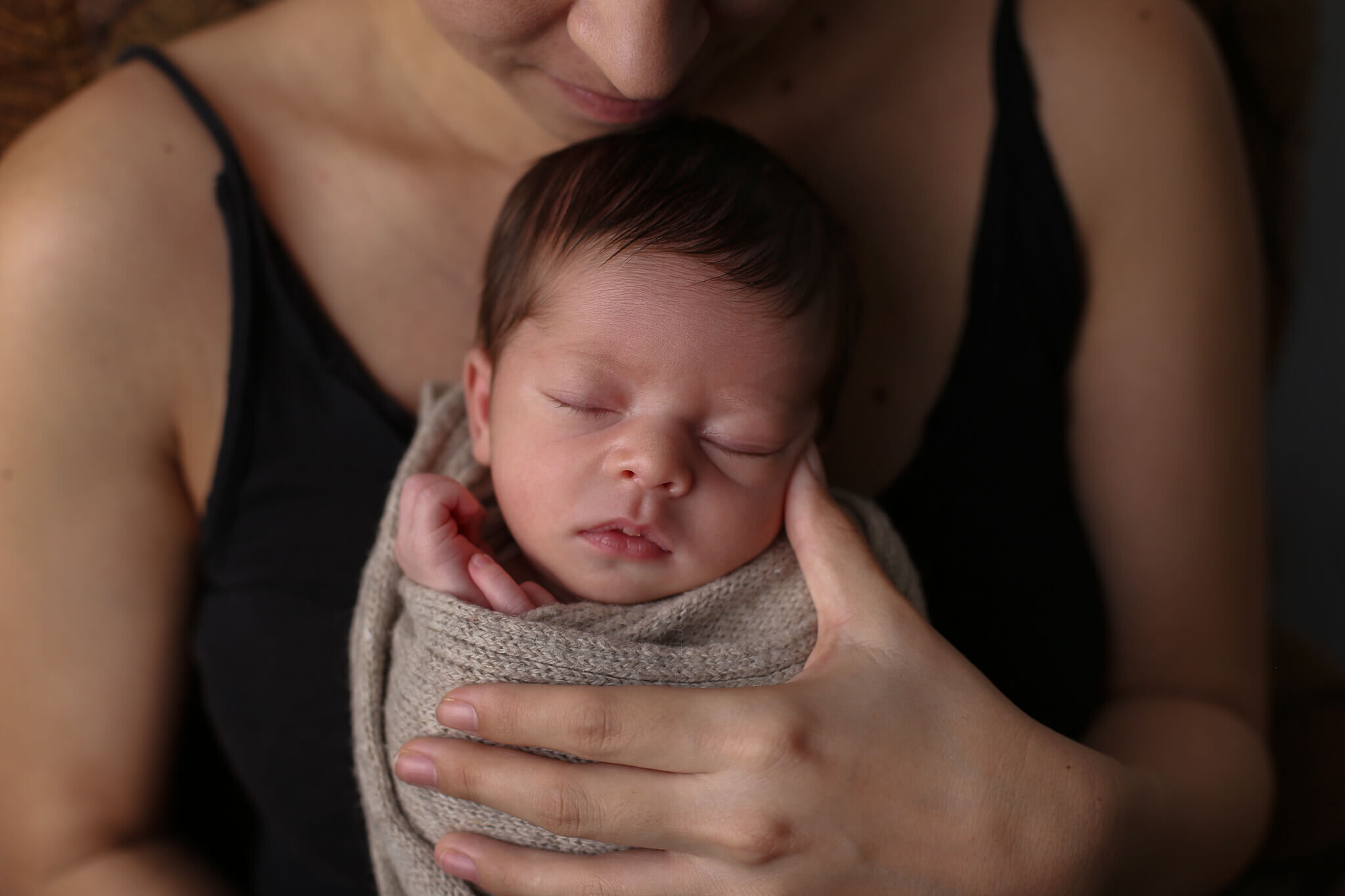  A picture of a peacefully sleeping newborn wrapped in a knitted blanket with its tiny hands peeking up near its face as mom holds the baby close to her chest from a newborn photo session 
