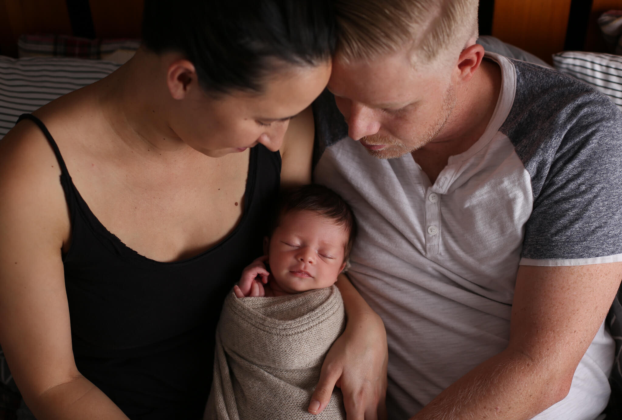  A photograph of a young family sitting close together as mom holds their newborn and dad gazes down as the baby sleeps by Photography by L Rose - San Diego CA newborn photography 