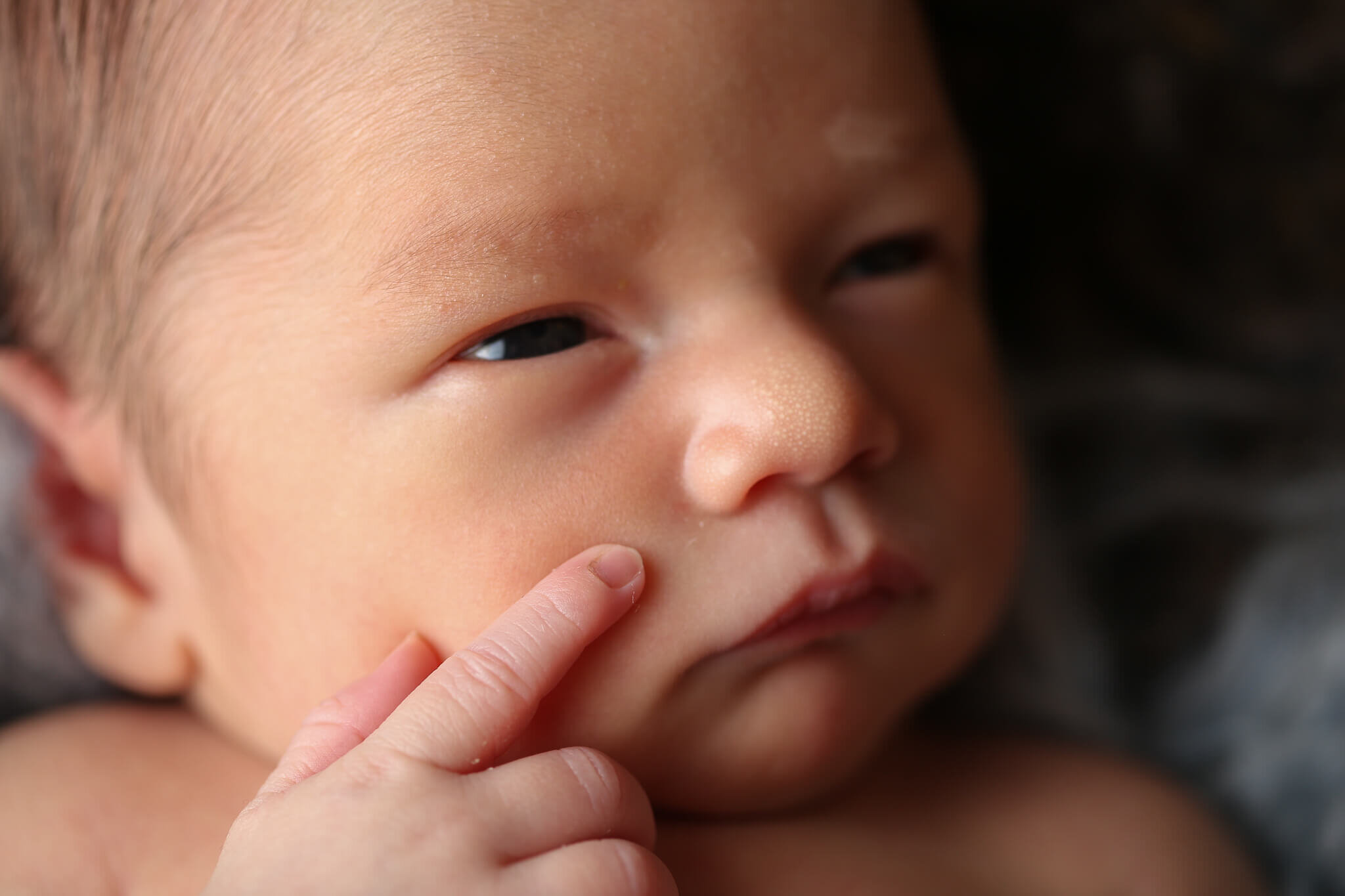  An image of a closeup view of a newborn baby’s face, eyes peering ahead in wonder as its tiny hand rests on its cheek, lit up in a warm glow of light from a newborn picture session 