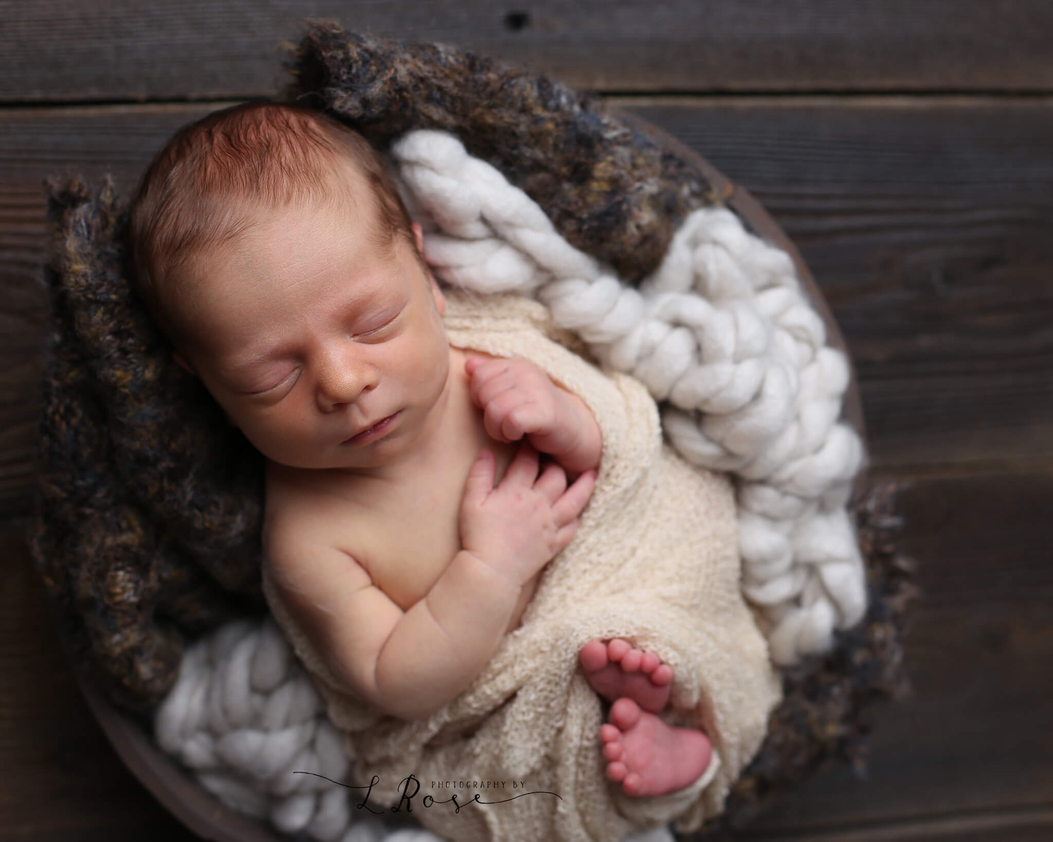  An image of a newborn baby wrapped tightly in gauze with hands and feet peeking out, sleeping in a basket lined with soft yarn blankets by Photography by L Rose in San Diego California 