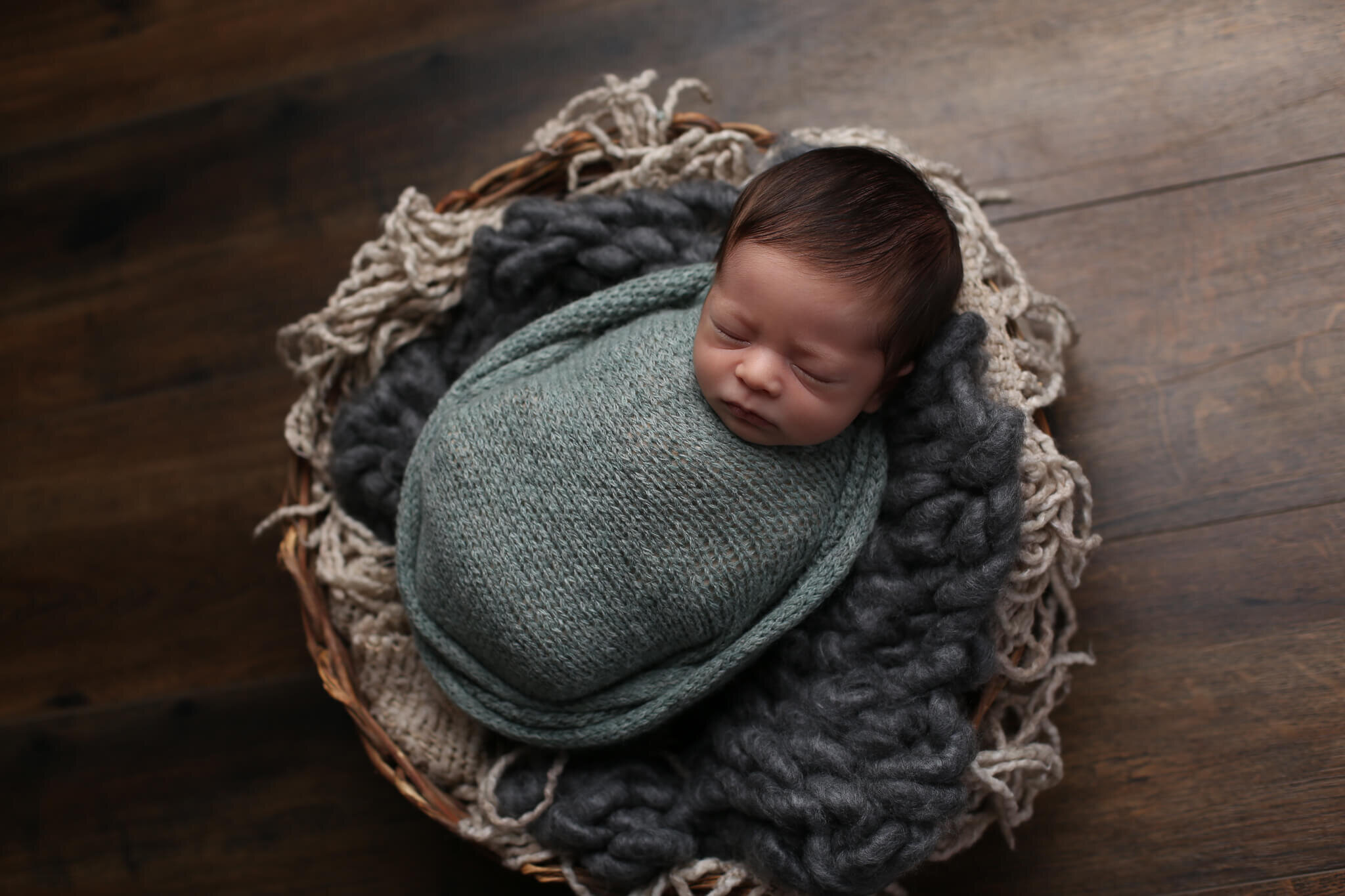  A photograph of a tiny newborn wrapped up in a knitted blanket, sleeping in a basket lined with cozy blankets from a baby photo session 