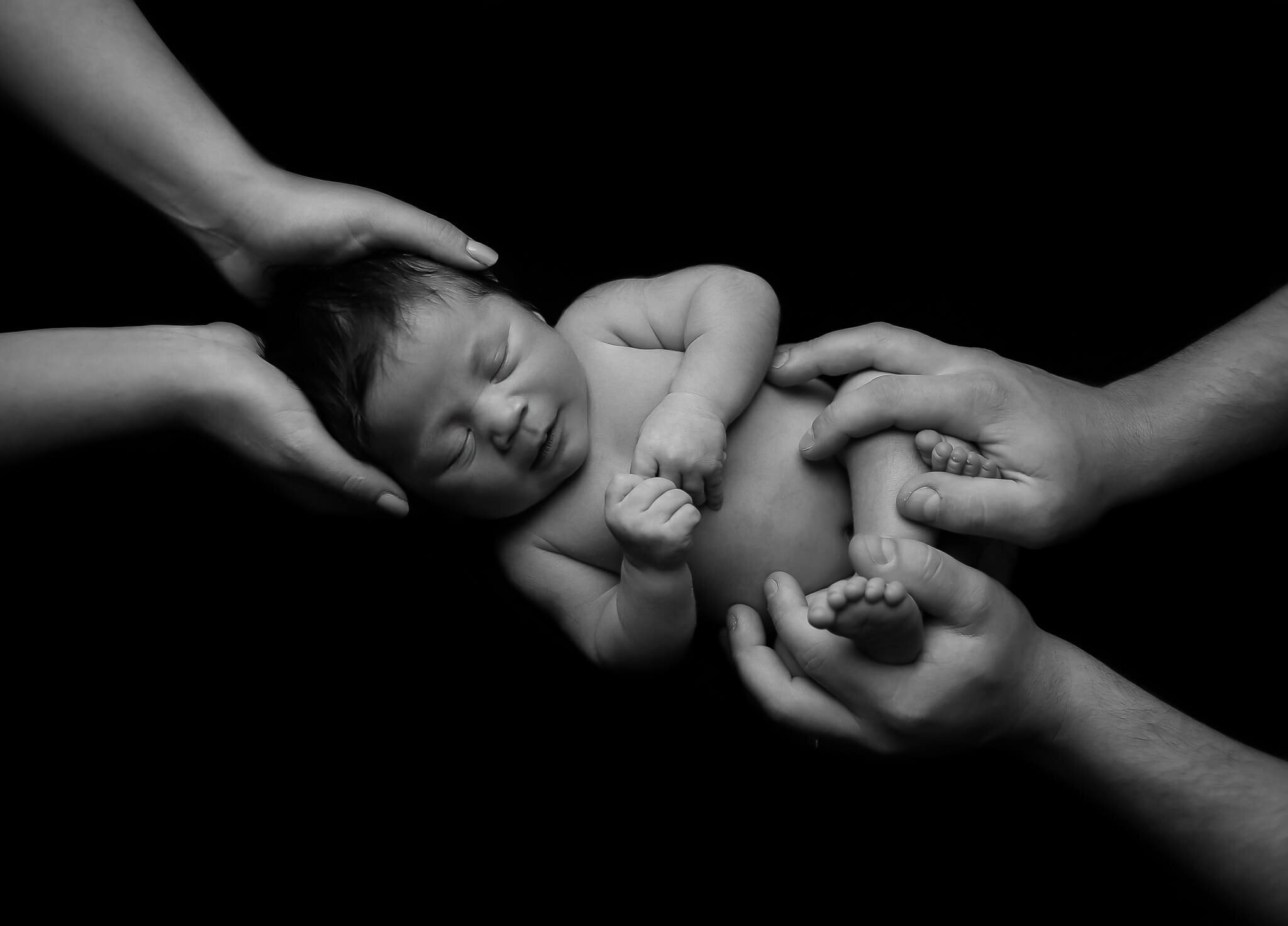  A picture in black and white of a tiny newborn sleeping with mom’s hands cradling the head and dad’s hands wrapped around the tiny legs from a newborn photo session 