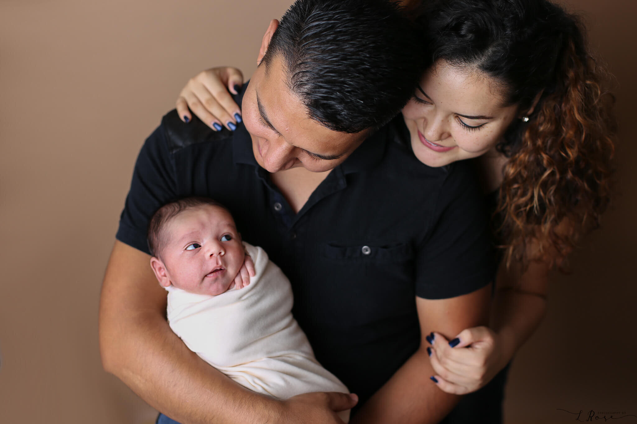  A photograph of a family with the new mother embracing the father from behind as he cradles the newborn in his arms and they gaze down at the wide awake baby from a baby photo session 