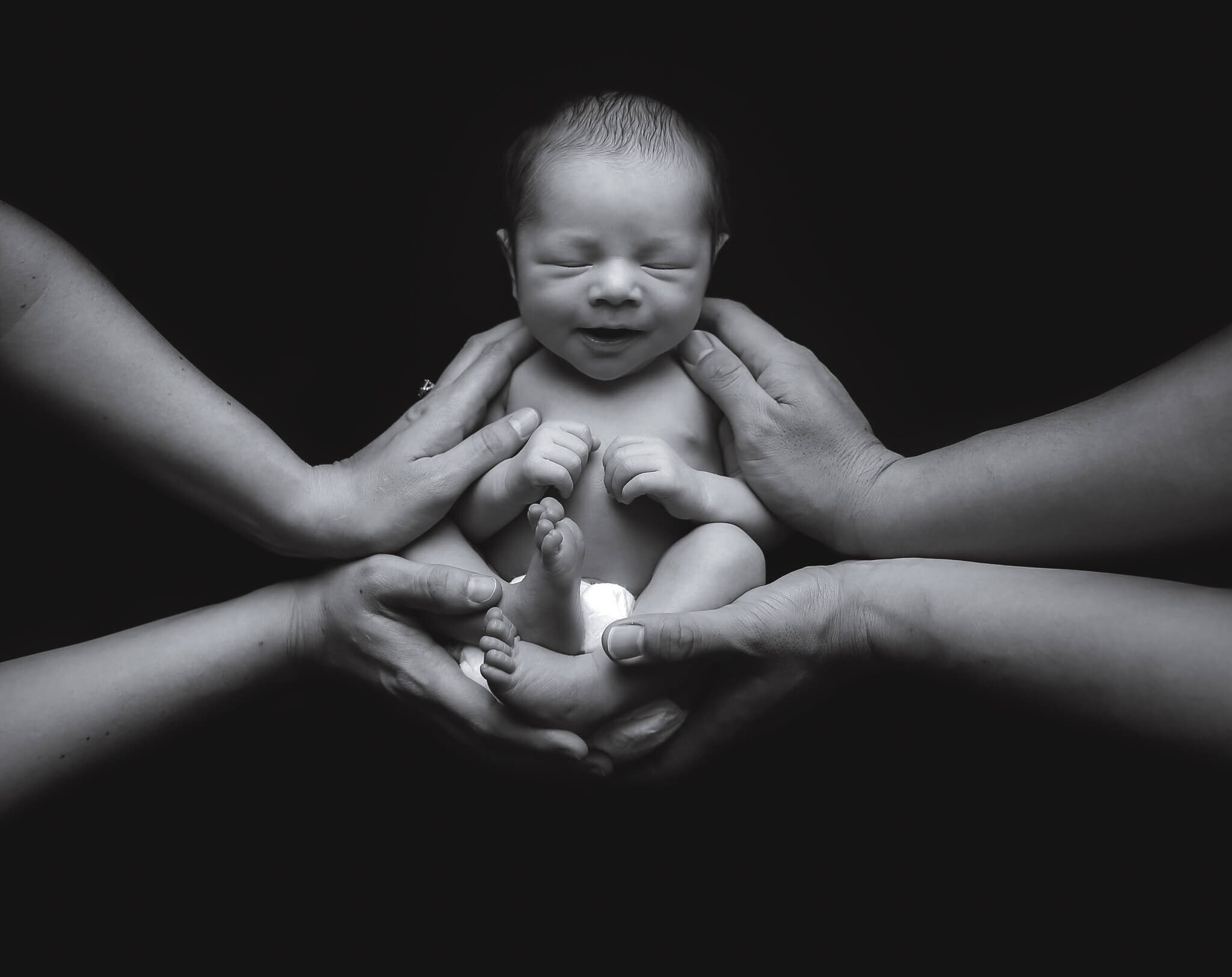  A picture in black and white of a tiny newborn lying down with his mother’s hands gently grasping his little body on one side while his father’s hands hold the other side 