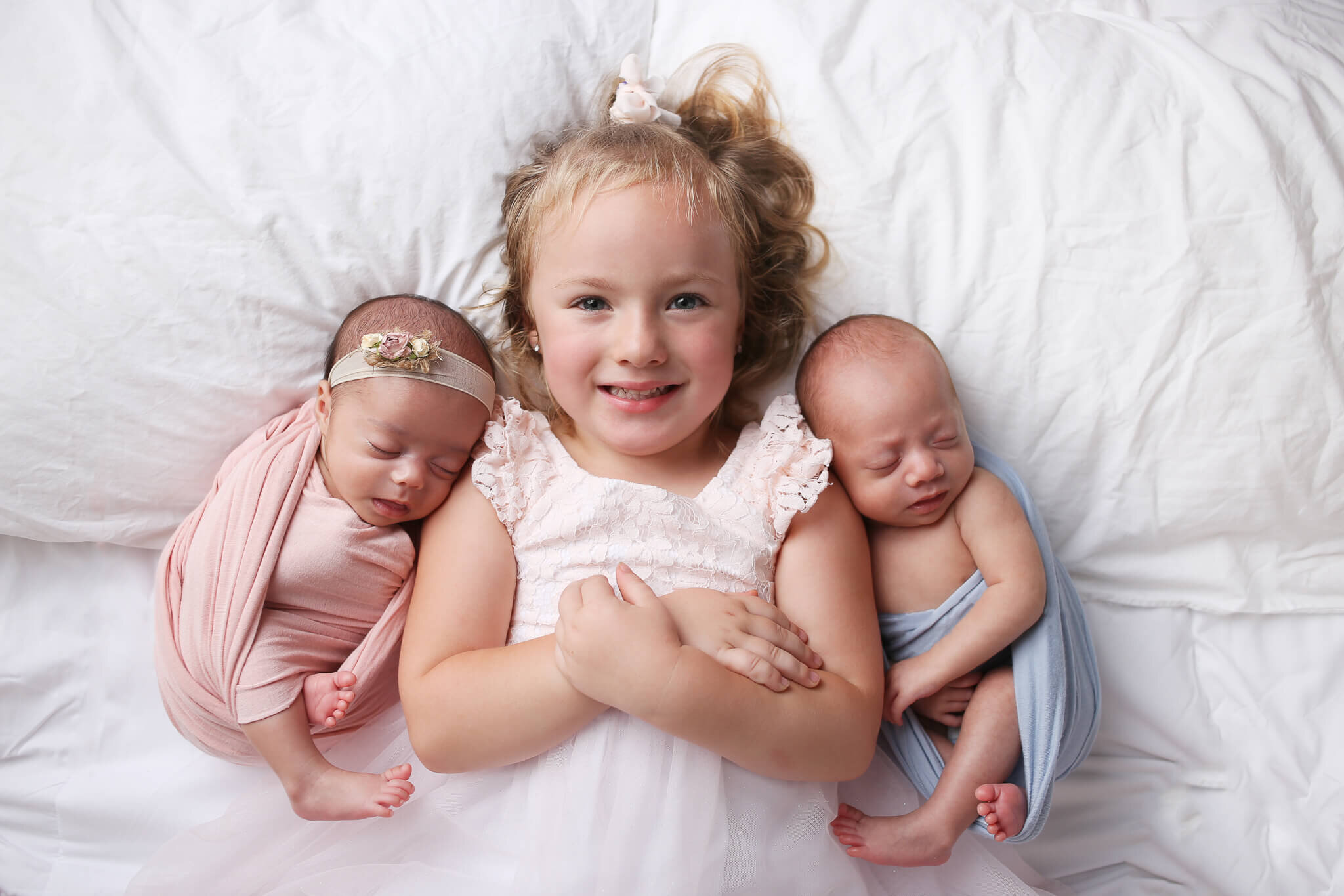  An image of two fraternal twin newborns, each swaddled in a tiny blanket, sleeping on either side of big sister as they lie in a bed side by side by Photography by L Rose 