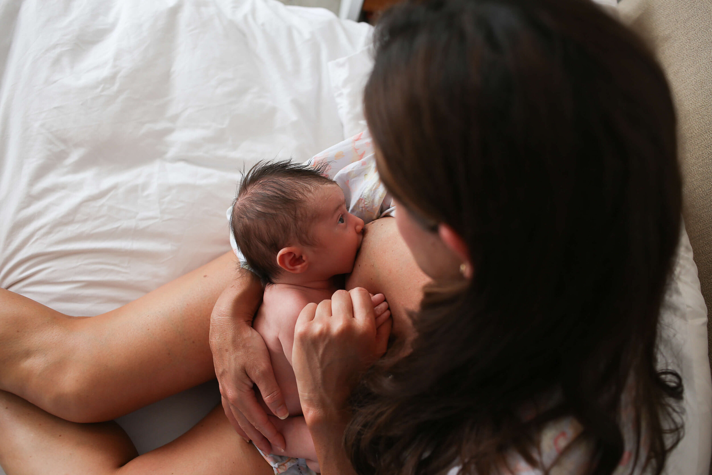  A picture from above of a tiny newborn nursing from mommy as they sit in bed with mom grasping his tiny hand, holding him close from a newborn photo session 