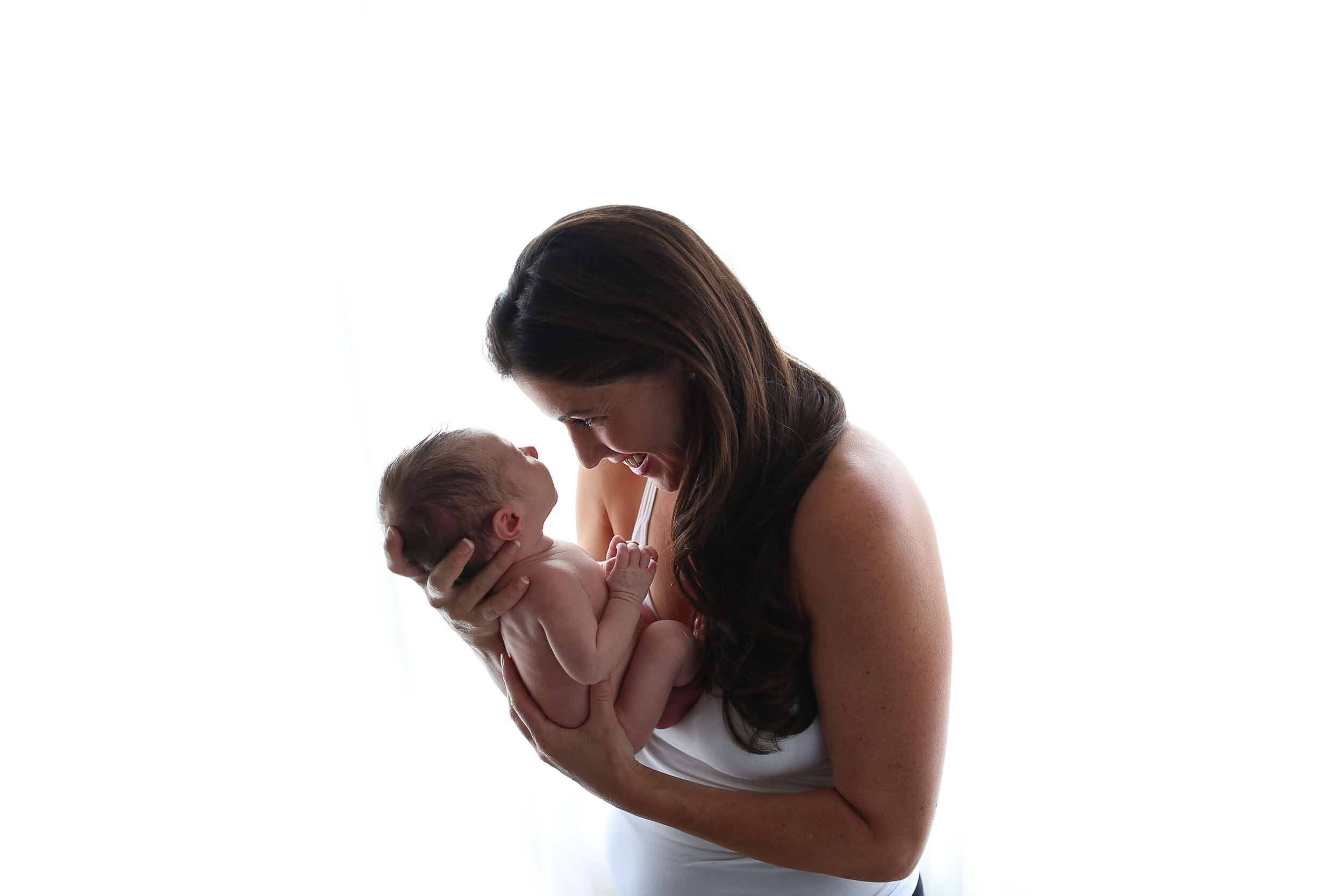  An image of a side view of a new mother holding her newborn baby up in front of her as she leans down to smile and talk to it, standing in front of a pure white background from a newborn picture session 