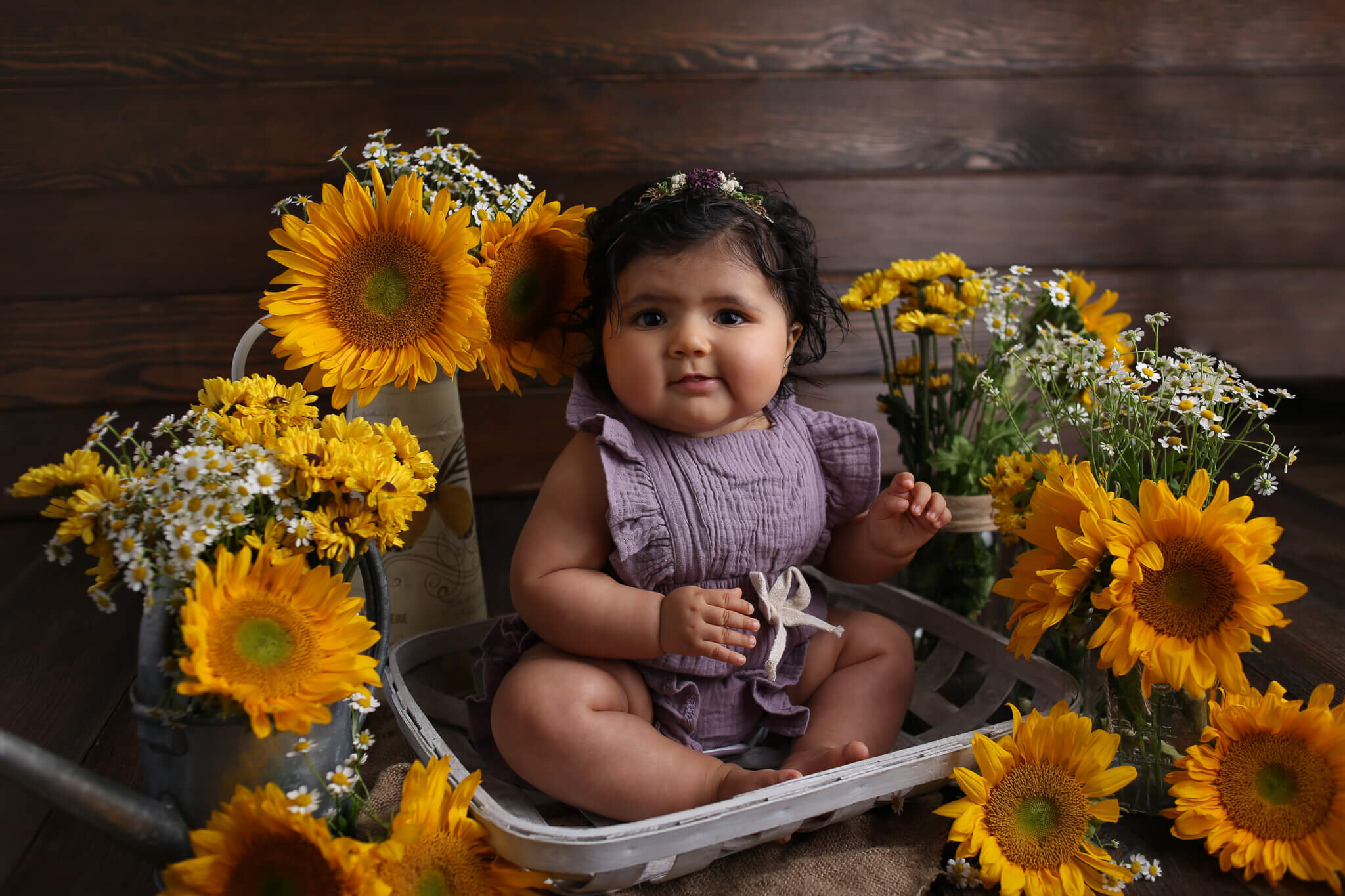  A photograph of a happy baby girl sitting in a little basket in her summer playsuit and headband with sunflowers and daisies in arrangements around her as she reaches a milestone by Photography by L Rose, a baby photographer in San Diego California 