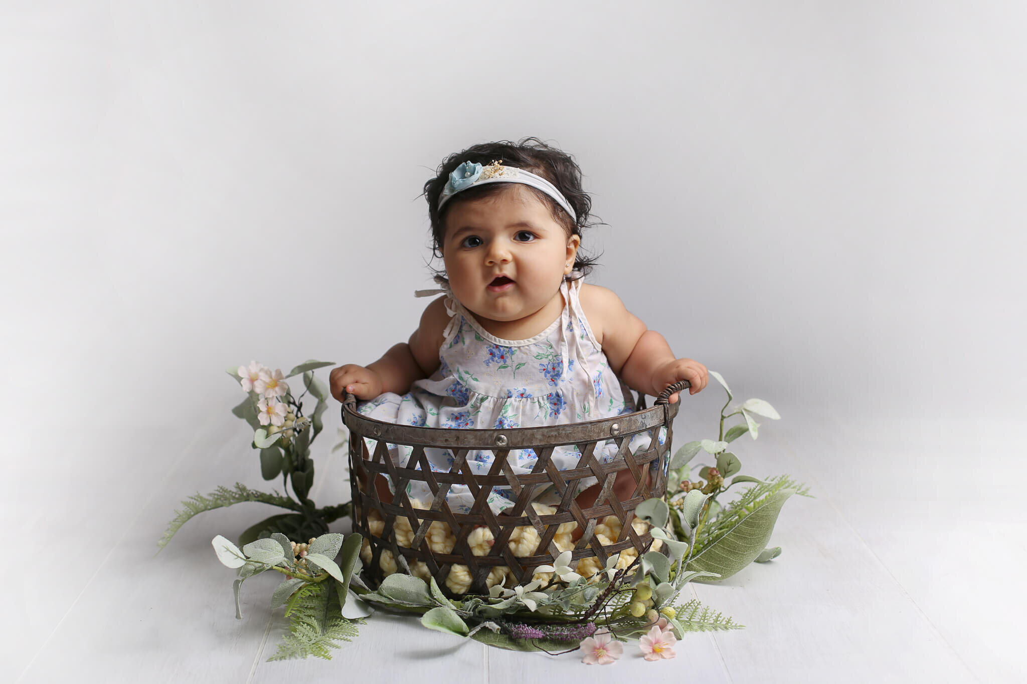  A picture of a sweet baby girl in a summery dress and headband sitting in a basket, surrounded by dainty flowers and graceful branches of leaves and ferns, celebrating a milestone by Photography by L Rose, baby photography in San Diego CA 