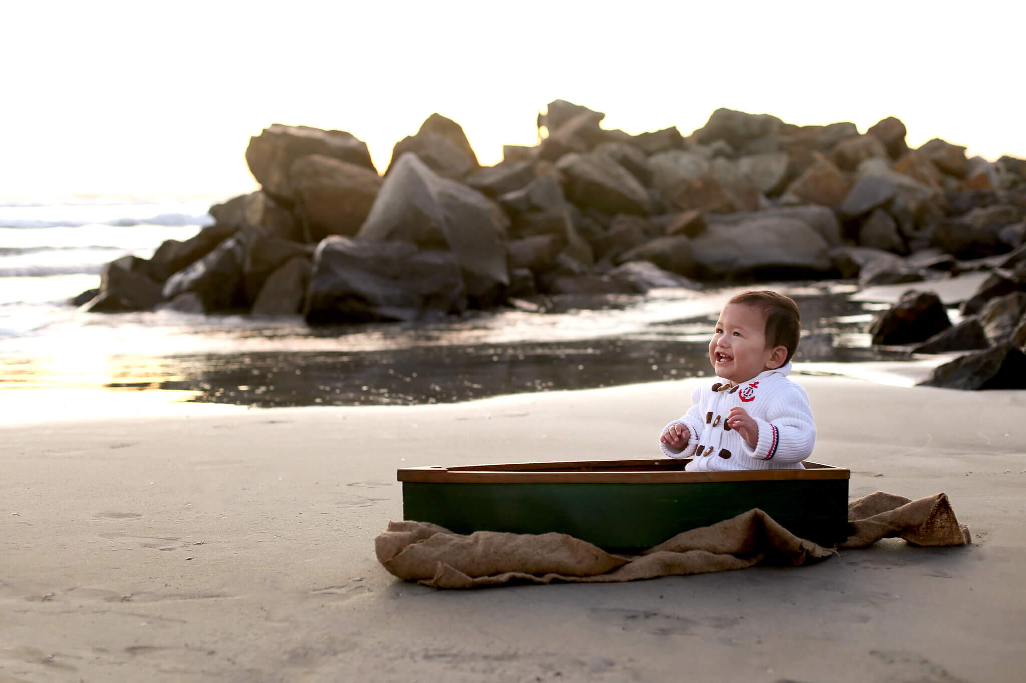  An image of a happy baby boy in a sporty sweater, sitting playfully in a tiny boat resting on the seashore near some rocks, marking his milestone from a baby milestone photo session 