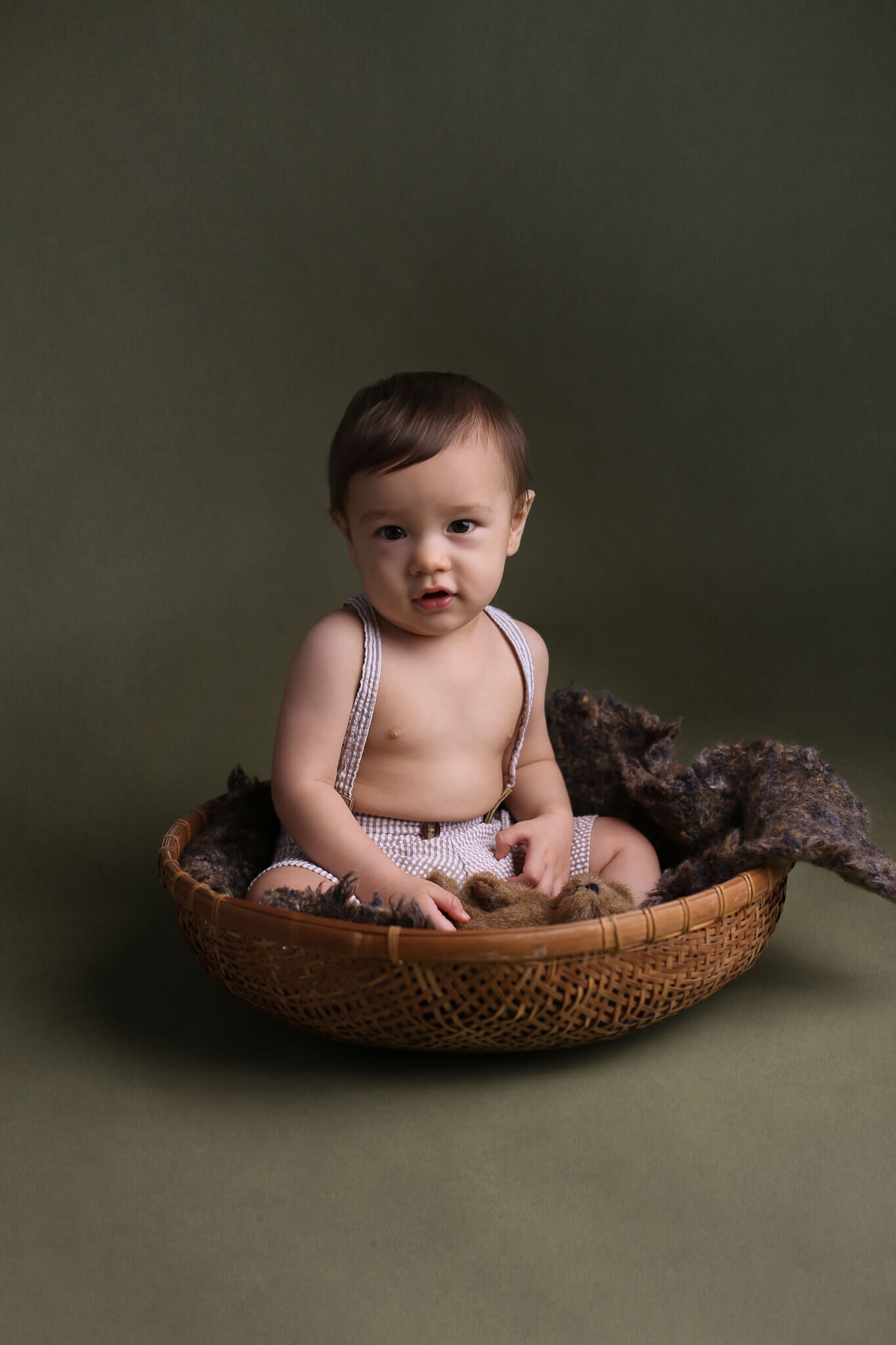  A picture of a baby boy sitting contentedly in his suspender shorts in a basket with a teddy bear in his hand, reaching a milestone in his young life from a baby photo session 