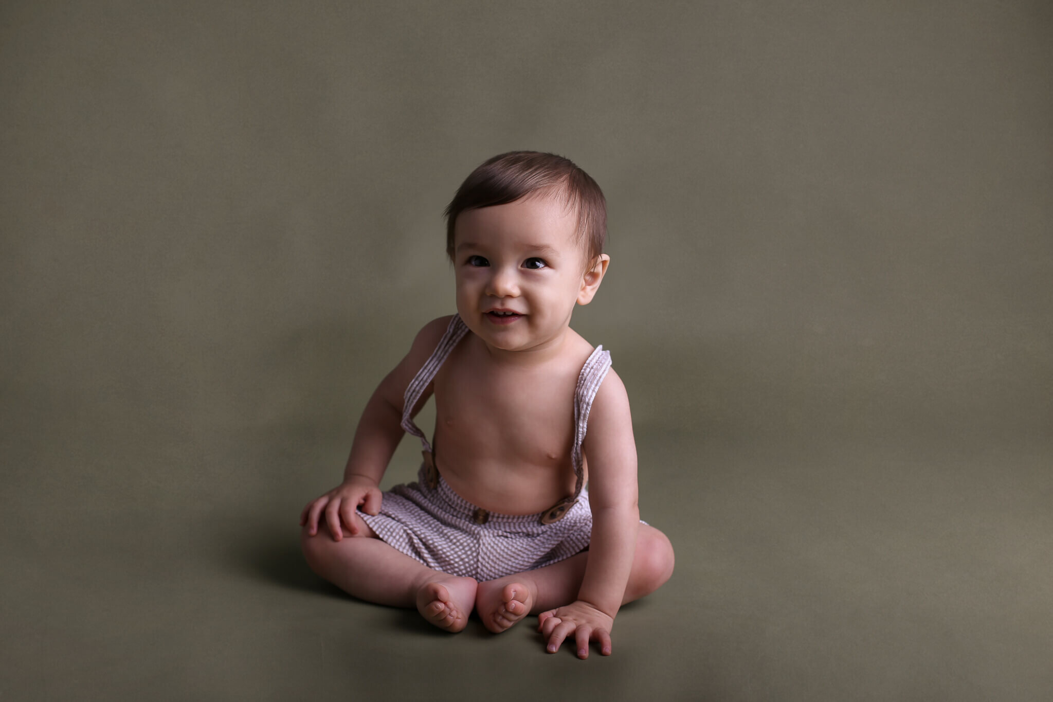  A photograph of a baby boy sitting in his cute suspender shorts, smiling and looking ahead with happy anticipation as he reaches a milestone from an infant picture session 