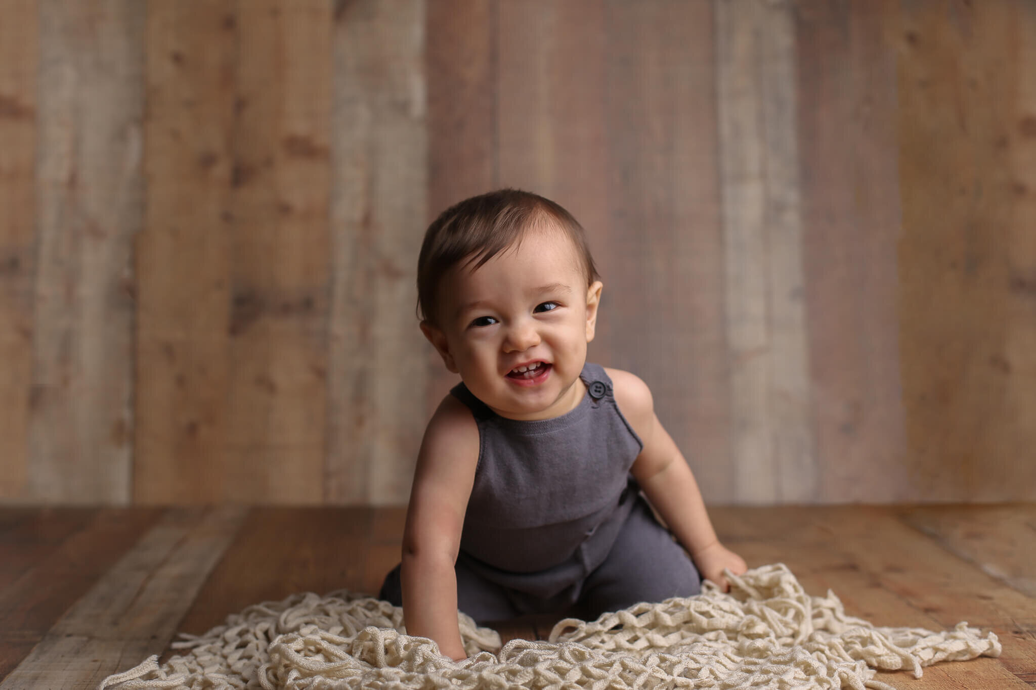  A photograph of a baby boy on his knees in a little jumpsuit, leaning forward and smiling playfully, showing his cute teeth as he celebrates a milestone by Photography by L Rose, a baby photographer in San Diego California 